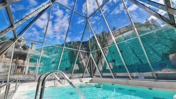 Una piscina interna con pareti di vetro trasparente, struttura ad intelaiatura metallica e vista su un paesaggio montuoso.