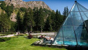 Uma estrutura em forma de pirâmide de vidro com uma piscina em primeiro plano, cercada por um jardim bem cuidado e um cenário montanhoso.
