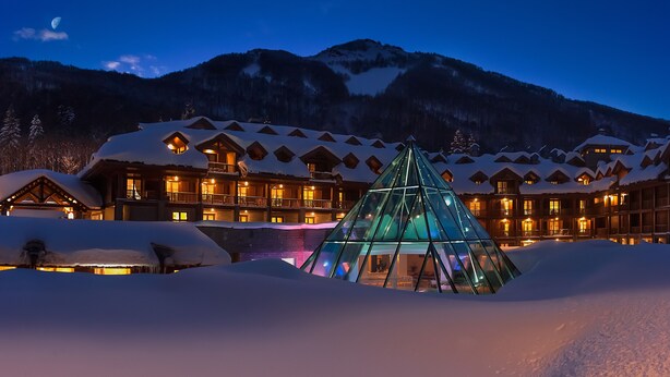 Uma pousada coberta de neve com vários edifícios, uma estrutura em forma de pirâmide de vidro e montanhas ao fundo.