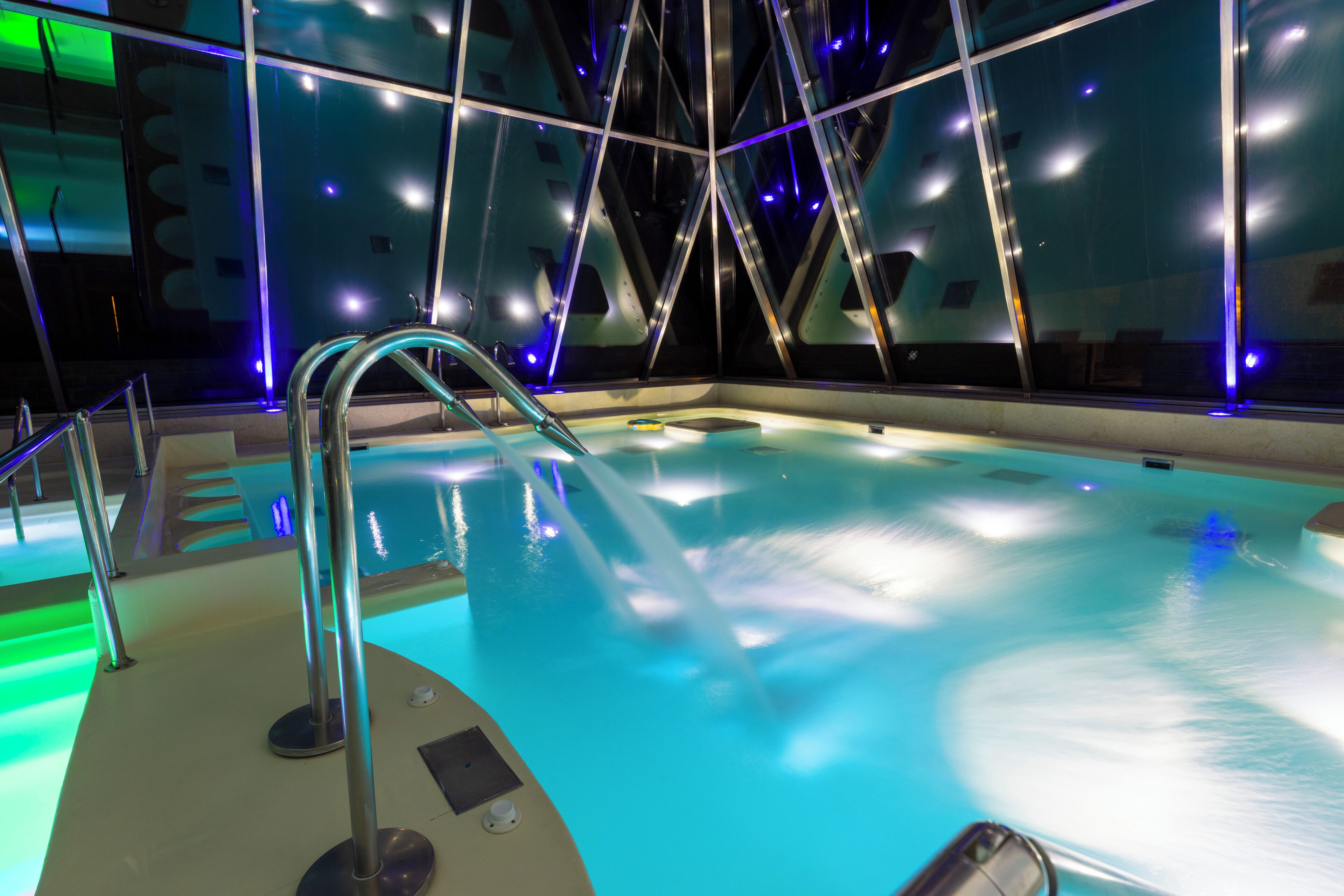 A modern indoor swimming pool with a glass wall, metal railings, and blue lighting.
