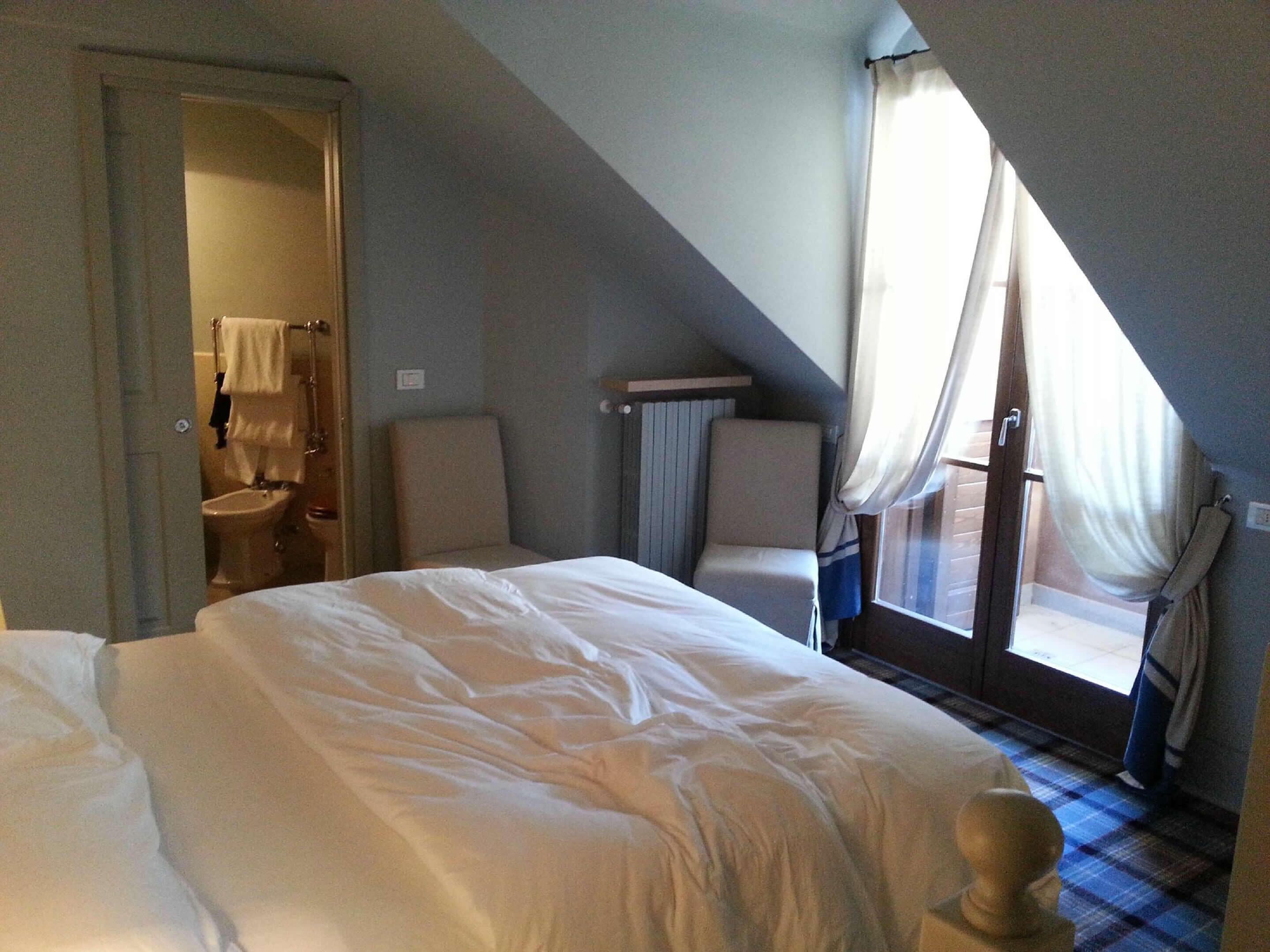 Une chambre à coucher avec un grand lit, une chaise, une fenêtre avec des rideaux et une porte donnant sur une salle de bain.