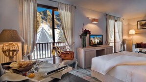 Quarto de hotel com uma cama, uma escrivaninha com televisão, uma janela com cortinas, um abajur e vista para as montanhas.