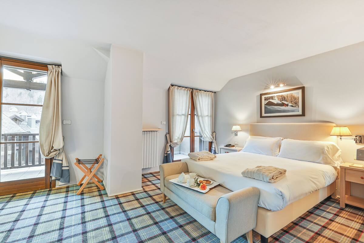 A hotel room with a large bed, a bedside table with a lamp, a window with curtains, a plaid-patterned floor, and a view of buildings outside.