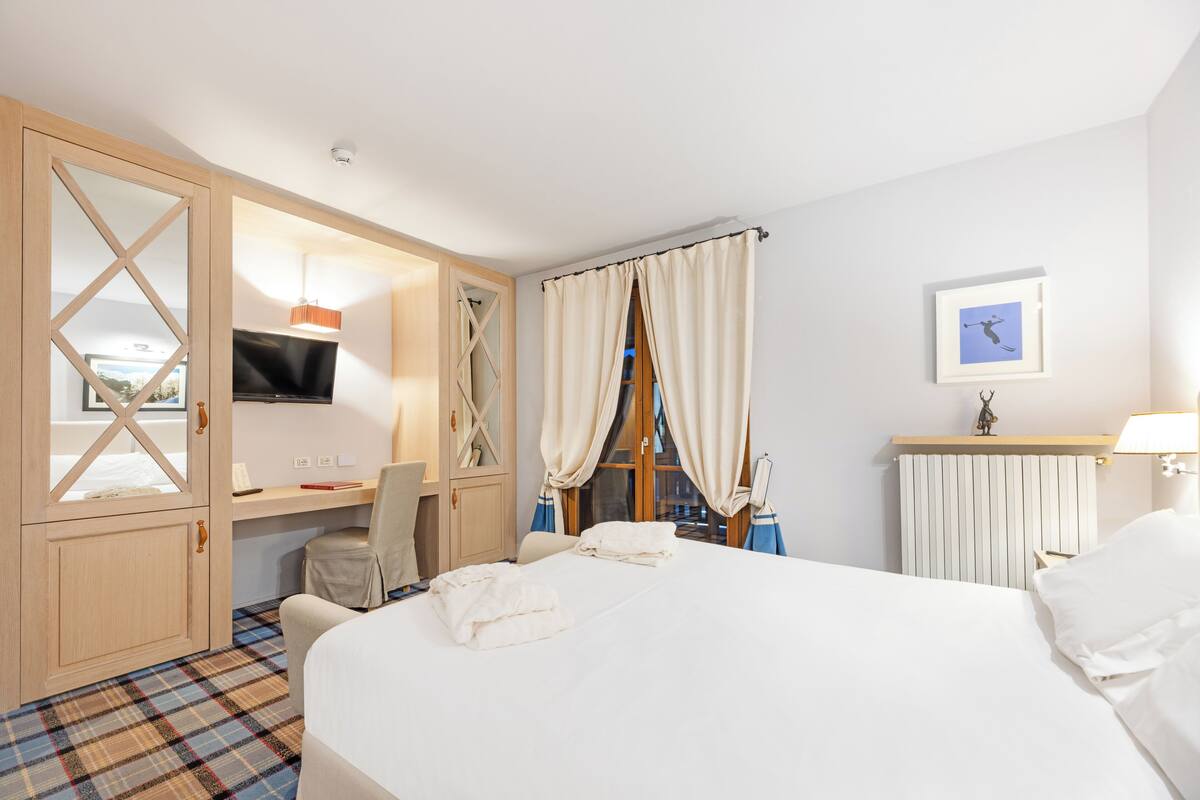 A hotel room with a bed, a desk with a chair, a television, and a window with curtains.