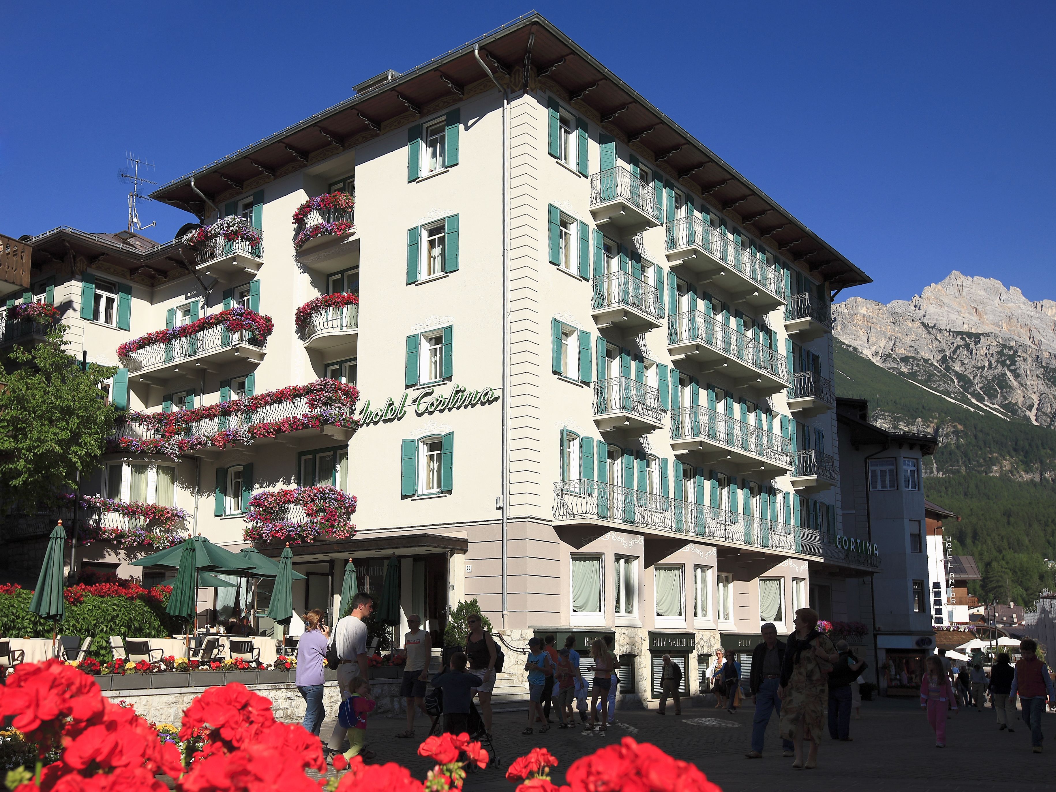A multi-story hotel with balconies adorned with flowers, a clear sky, and a mountainous backdrop.