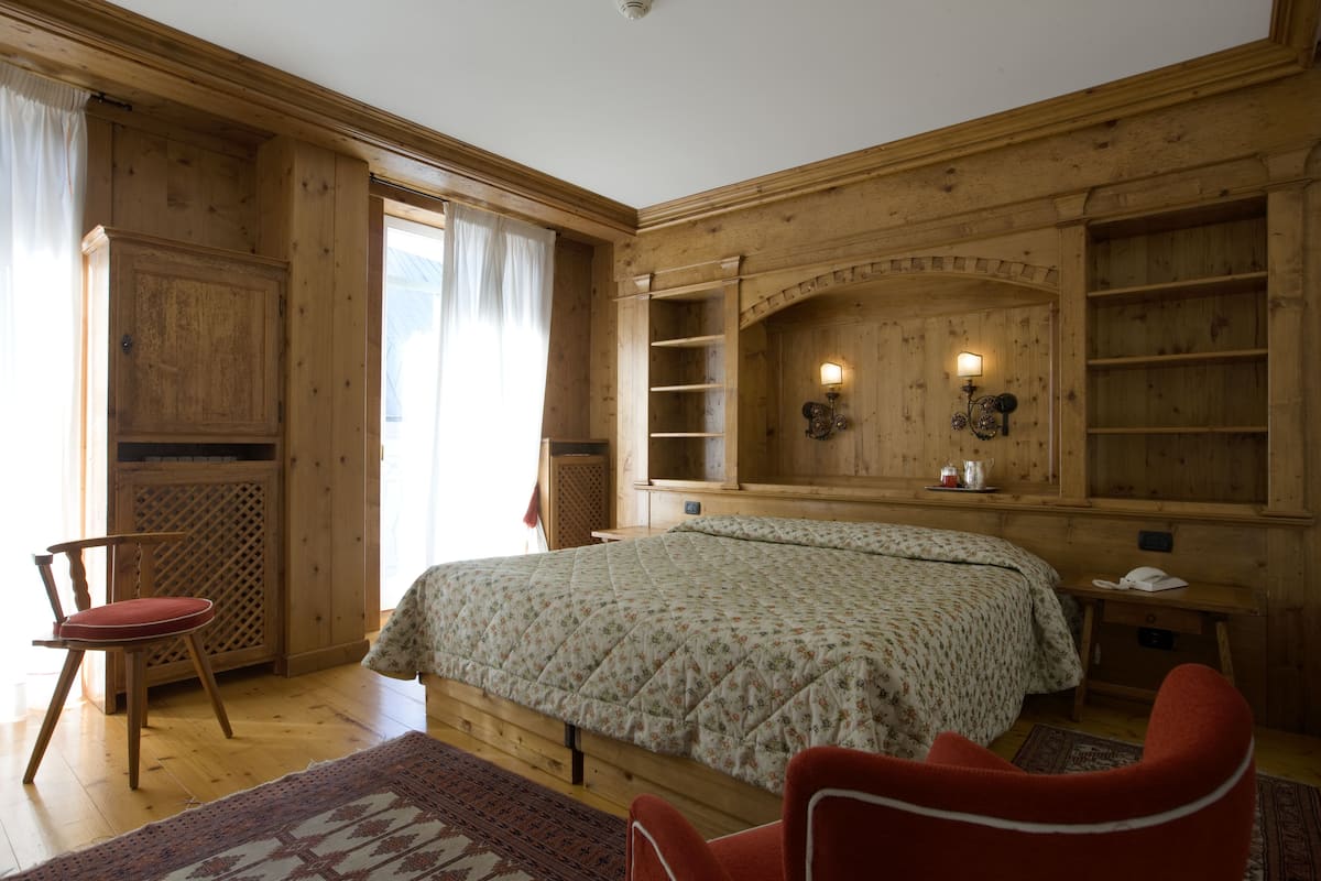 A wooden-paneled bedroom with a bed, a chair, a nightstand, and a shelf with a lamp.