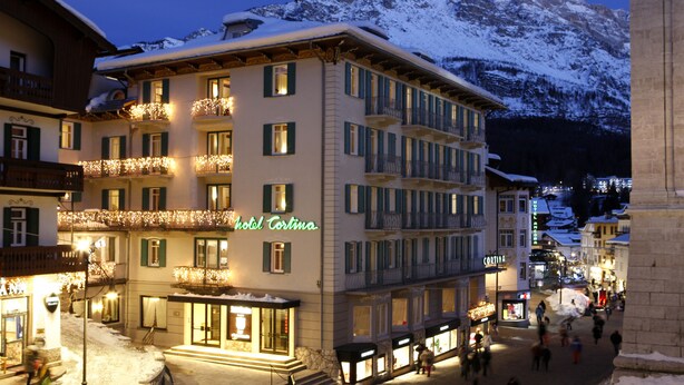 A hotel with multiple balconies, a snow-covered mountain in the background, and a street with people walking. - Cortina (Garda)