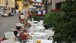 Outdoor dining tables with white tablecloths and floral centerpieces, surrounded by greenery and a cobblestone street. - Cortina (Garda)