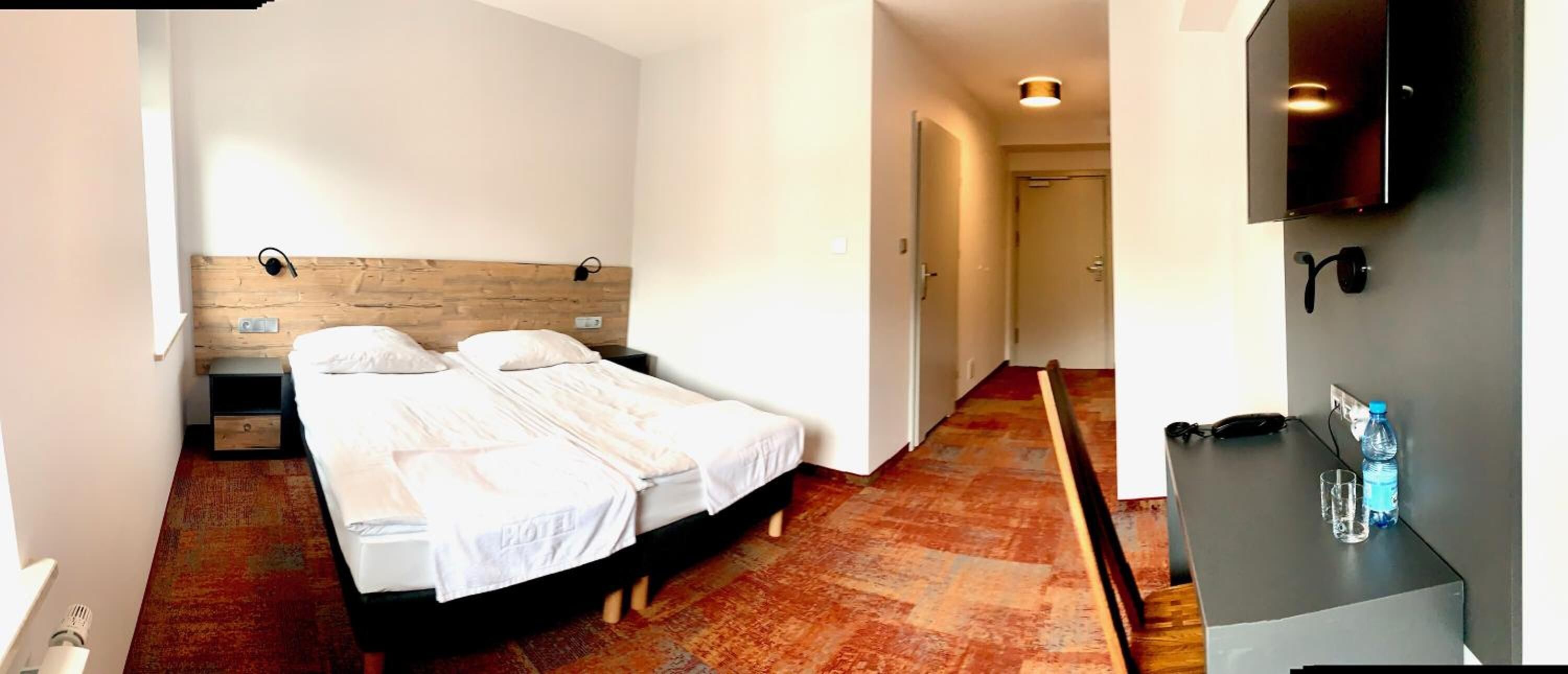 A hotel room with a wooden headboard bed, a desk with a chair, and a flat-screen TV mounted on the wall.