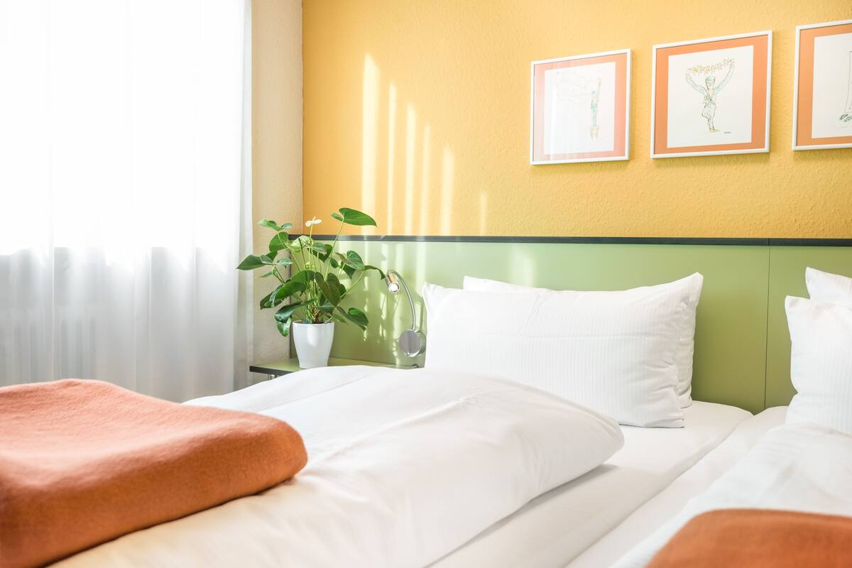 Quarto de hotel com uma cama, cabeceira verde, roupa de cama branca, uma planta em vaso e obras de arte emolduradas na parede.