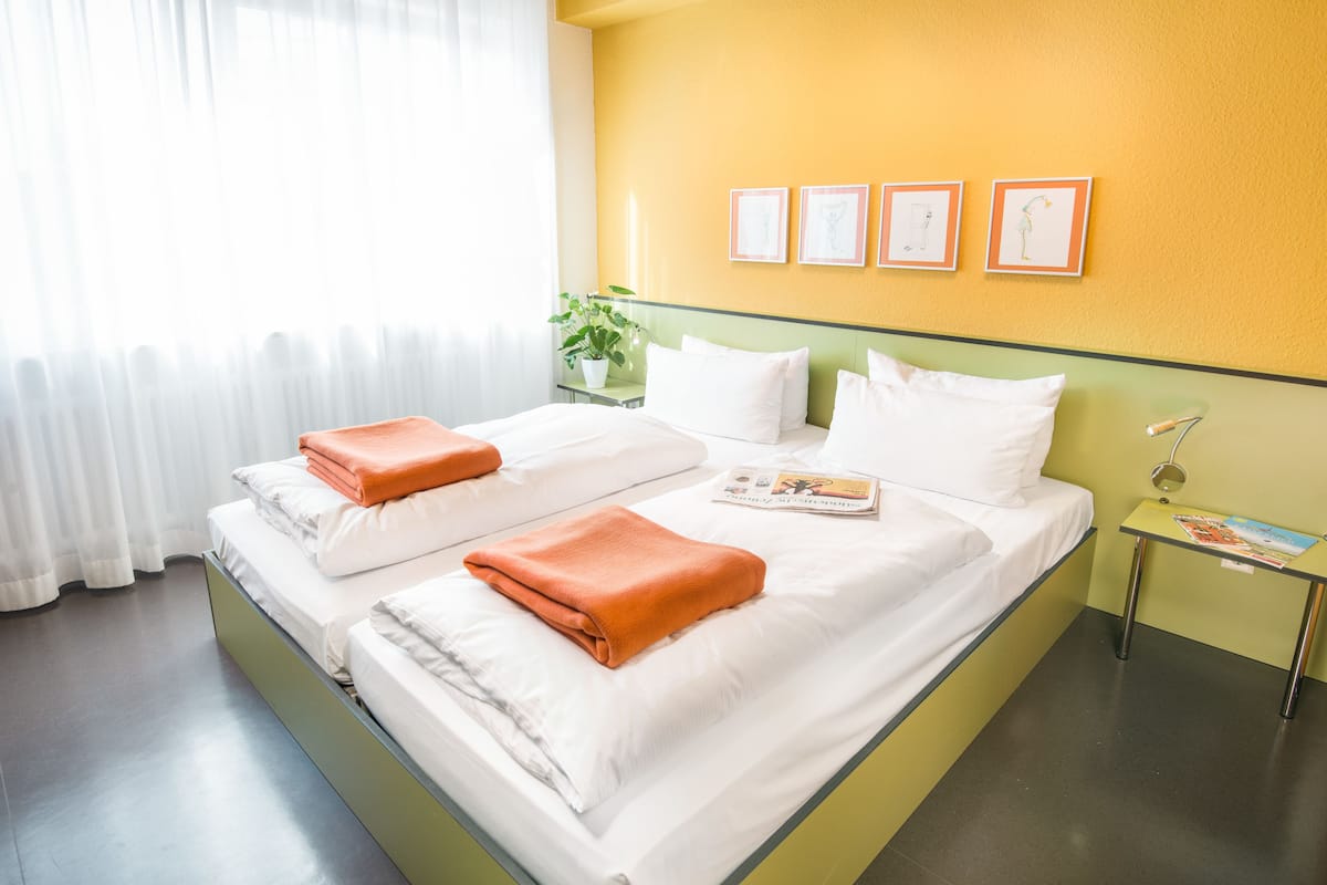 Cama de casal com roupa de cama branca e toalhas laranja, mesa de cabeceira com abajur e janela com cortinas translúcidas.