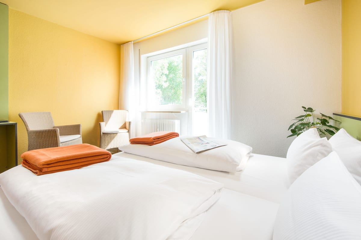 Quarto de hotel com duas camas, uma cadeira, uma mesa e uma janela com cortinas.