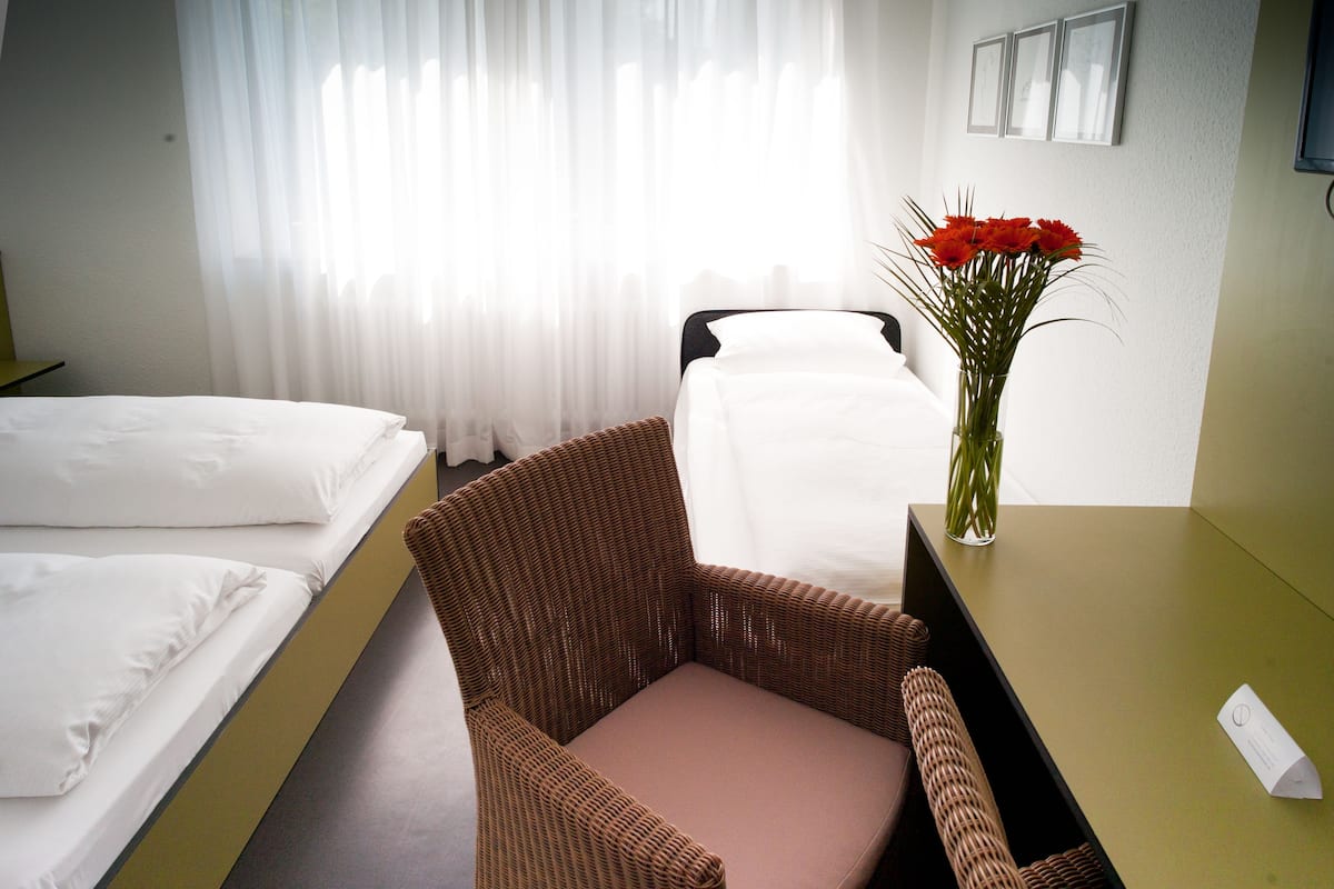 Quarto de hotel com cama, uma cadeira de vime, uma mesa pequena e um vaso com flores.