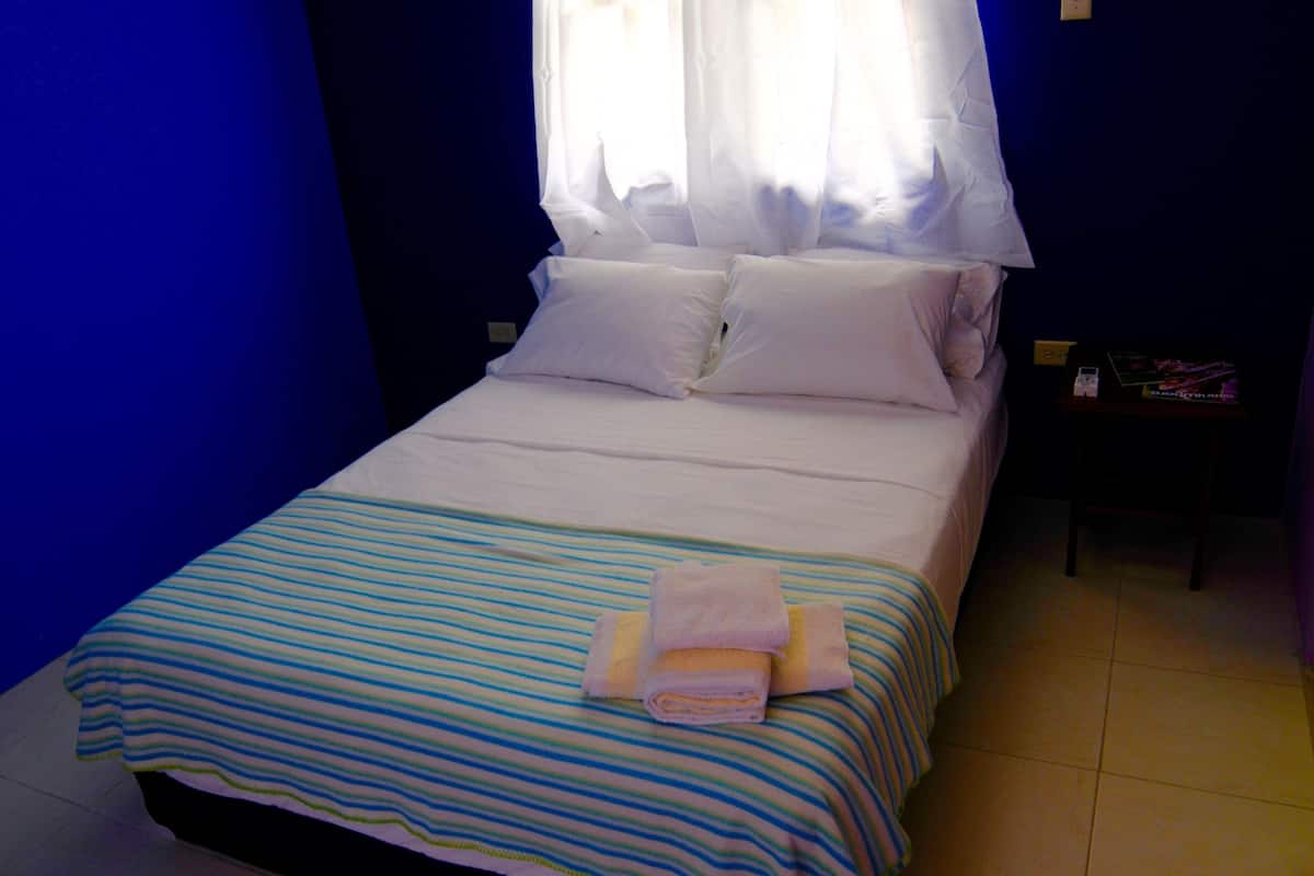 A neatly made bed with a striped bedspread and a folded towel, a nightstand with a book and a phone, and a window with sheer curtains.