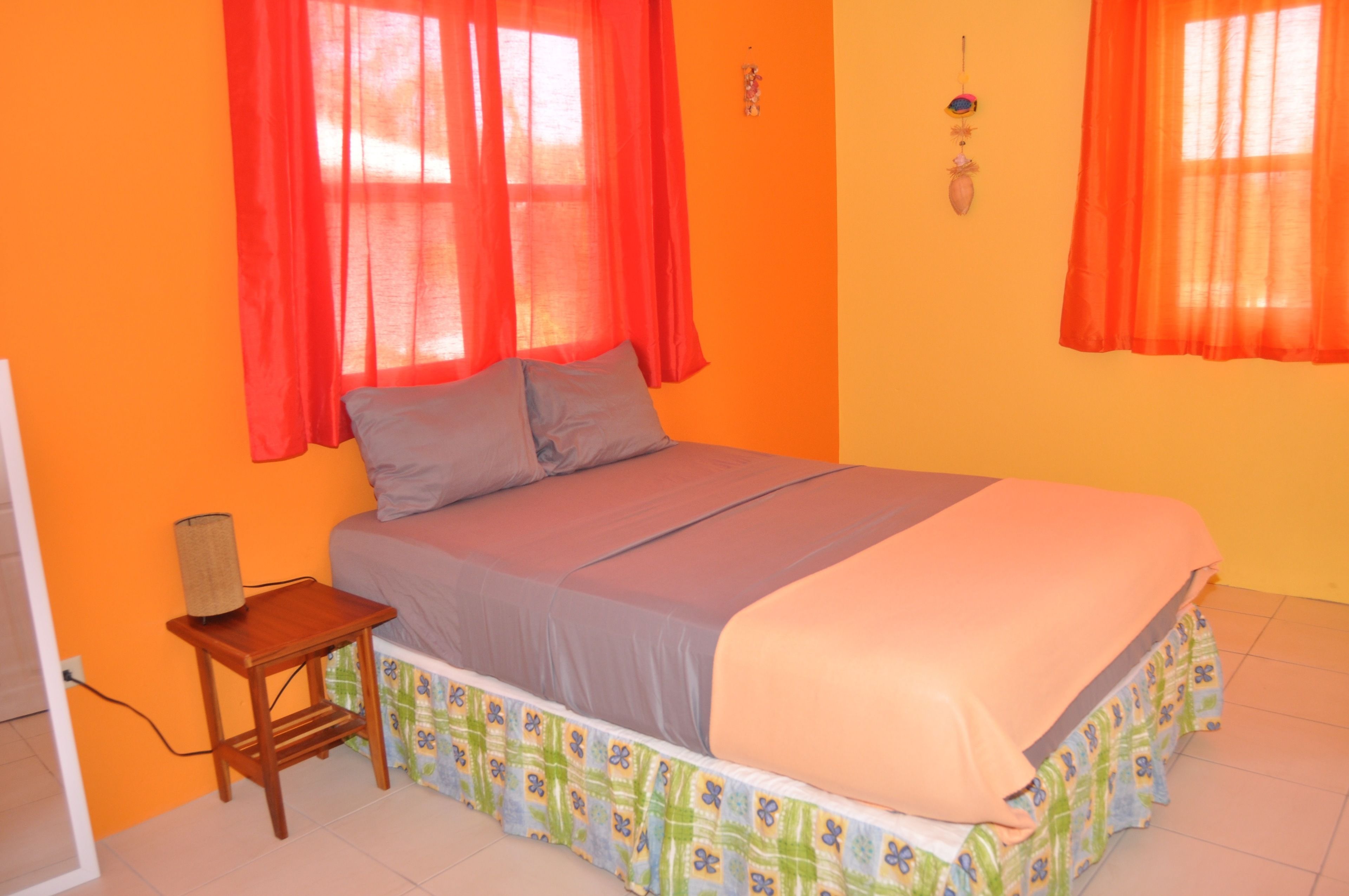 A bedroom with a bed, two pillows, a bedside table with a lamp, and two windows with red curtains.