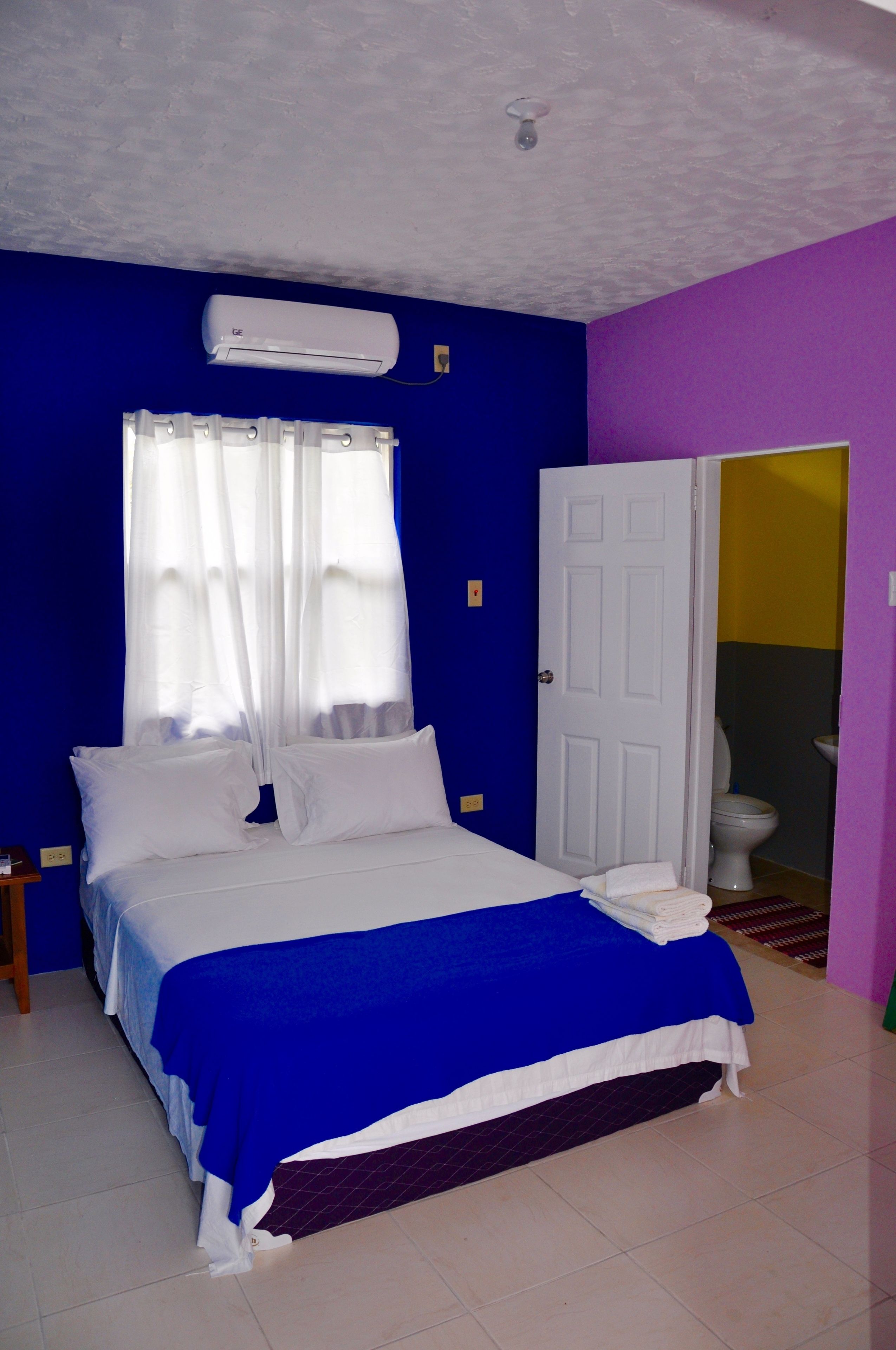 Un dormitorio con cama, cortinas blancas, una puerta blanca y un baño visible a través de la puerta.