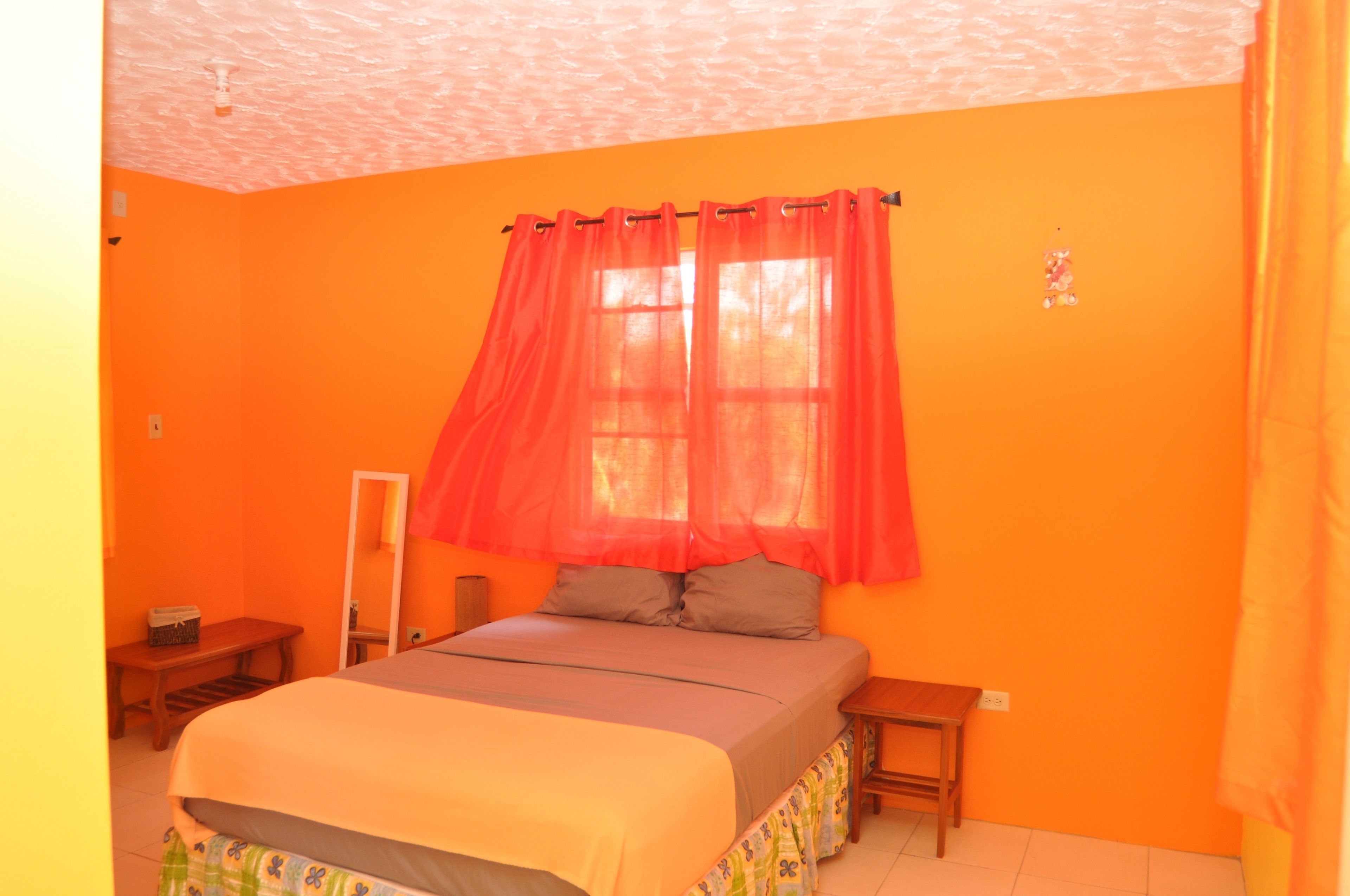A bedroom with a bed, a nightstand, a mirror, and a window with red curtains.