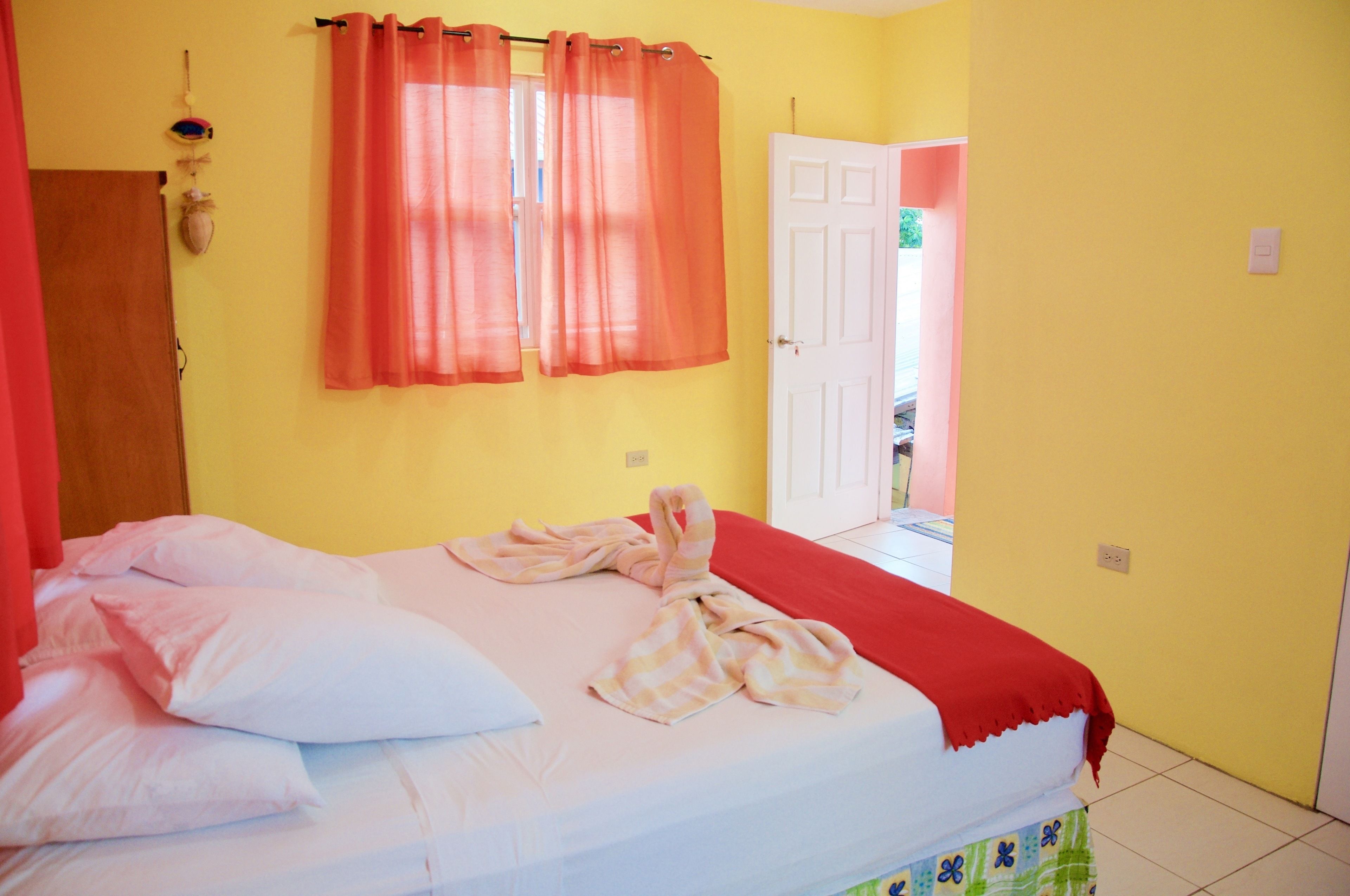 A bedroom with a bed, pillows, a red blanket, and a window with orange curtains.