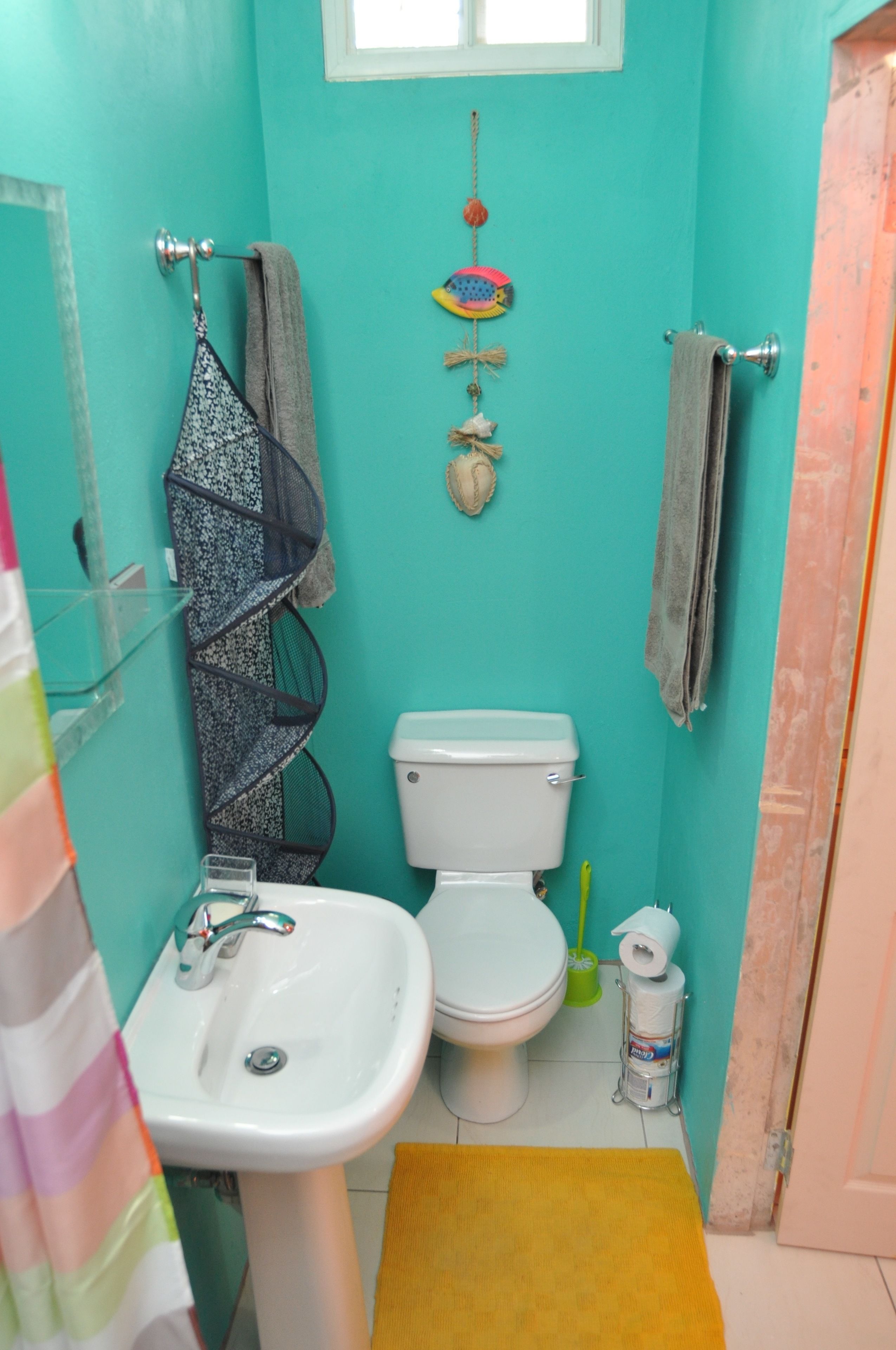 Une petite salle de bain avec des toilettes, un lavabo et un tapis jaune. Des serviettes sont accrochées au mur, et une petite étagère contient des articles de toilette.
