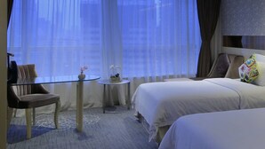 Une chambre d’hôtel avec deux lits, une petite table ronde ornée d’un vase de fleurs, une chaise et une grande fenêtre avec des rideaux légers.
