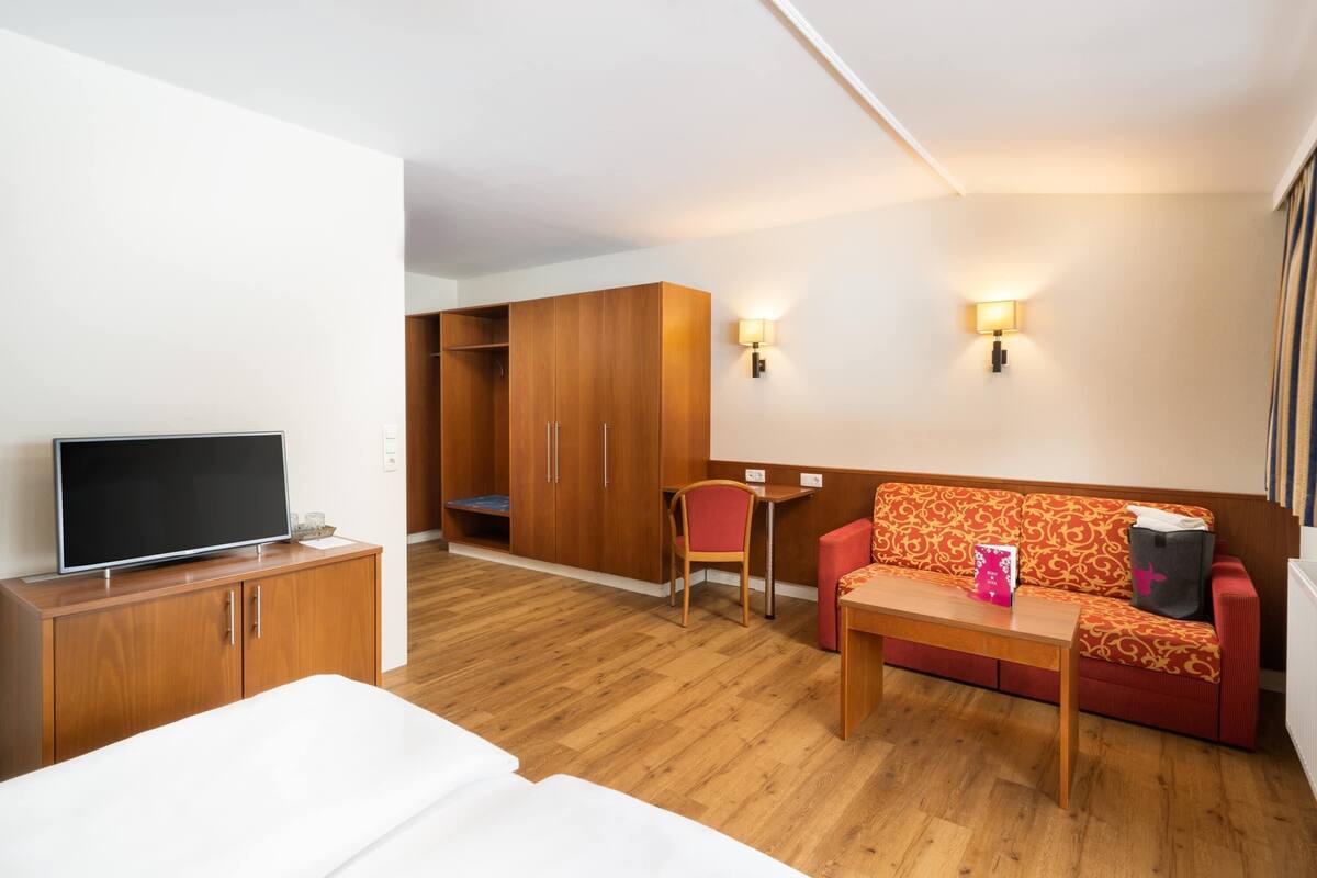 Une chambre d’hôtel avec une armoire en bois, un téléviseur posé sur un meuble, un canapé rouge et une petite table.