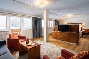 A modern hotel room with a sofa, a TV, a wooden cabinet, and a dining table with fruit.