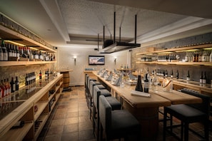 A bar area with multiple shelves of bottles, a long counter with bar stools, and a television screen on the wall.