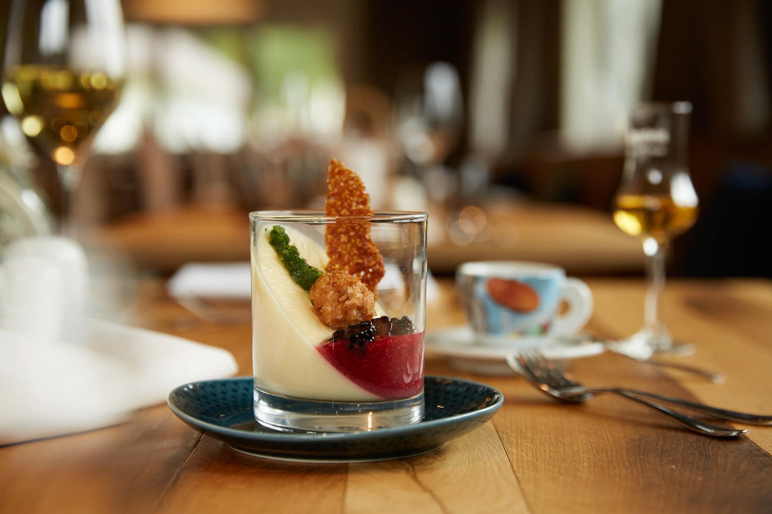 Ein Dessert im Glas auf einem blauen Teller, mit einem Löffel und einer Tasse im Hintergrund.