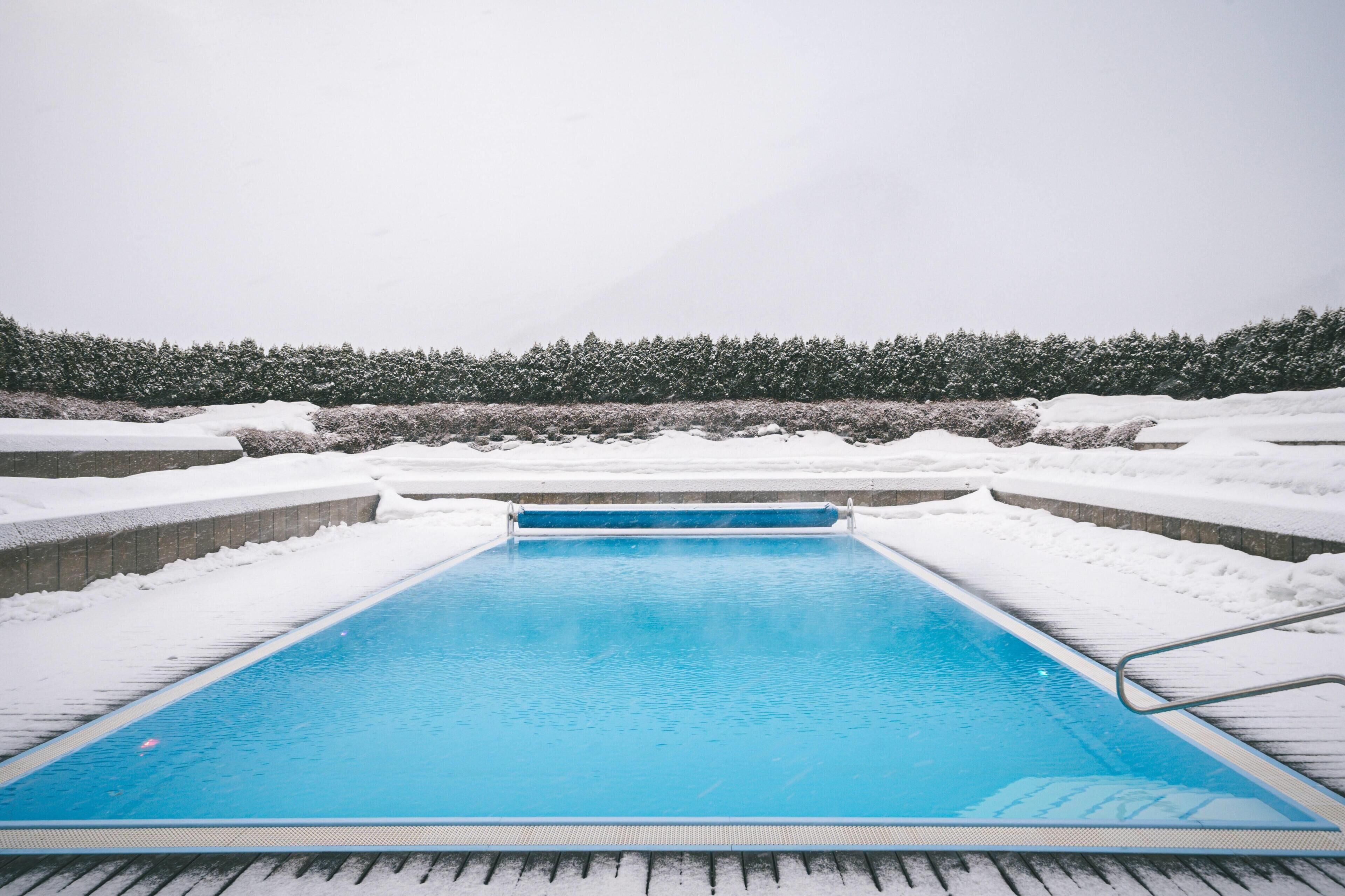 A rectangular swimming pool with a blue interior, surrounded by snow-covered ground and a hedge in the background.