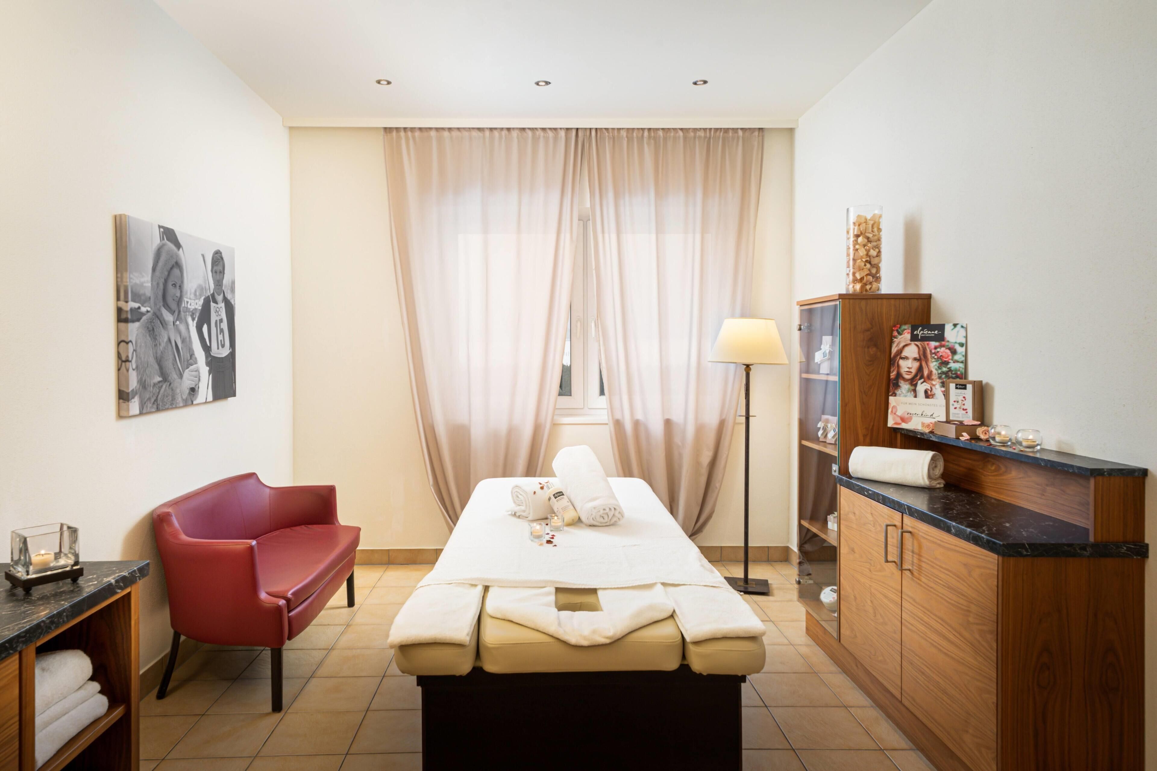 A room with a massage table, a red chair, a cabinet with toiletries, and a large window with curtains.