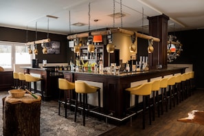 Una zona de bar con una barra alargada, varios taburetes, luces colgantes y una gran variedad de botellas y vasos.