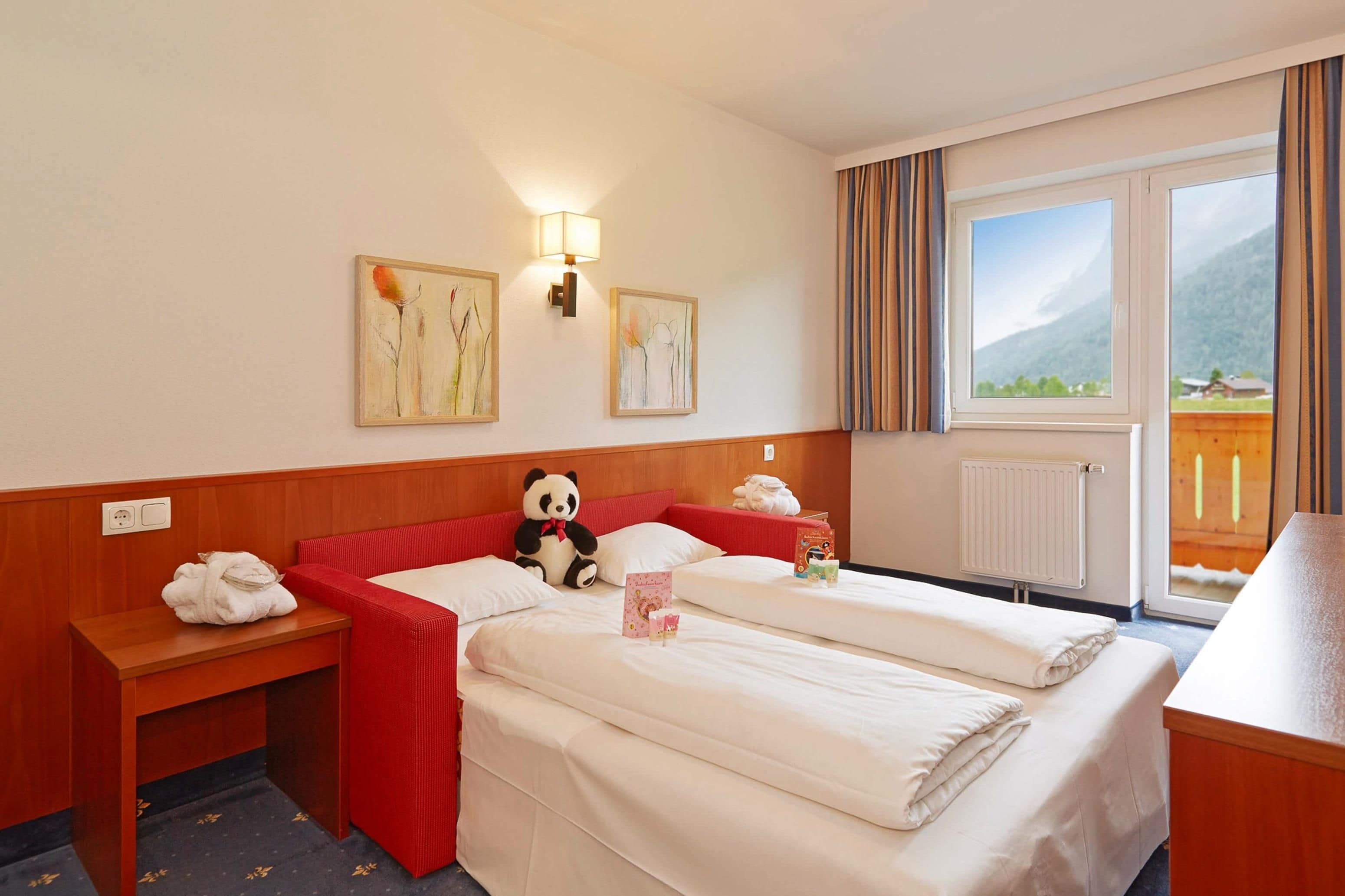 Habitación de hotel con una cama, una mesita de noche con una toalla, un ventanal con cortinas y dos cuadros colgados en la pared.