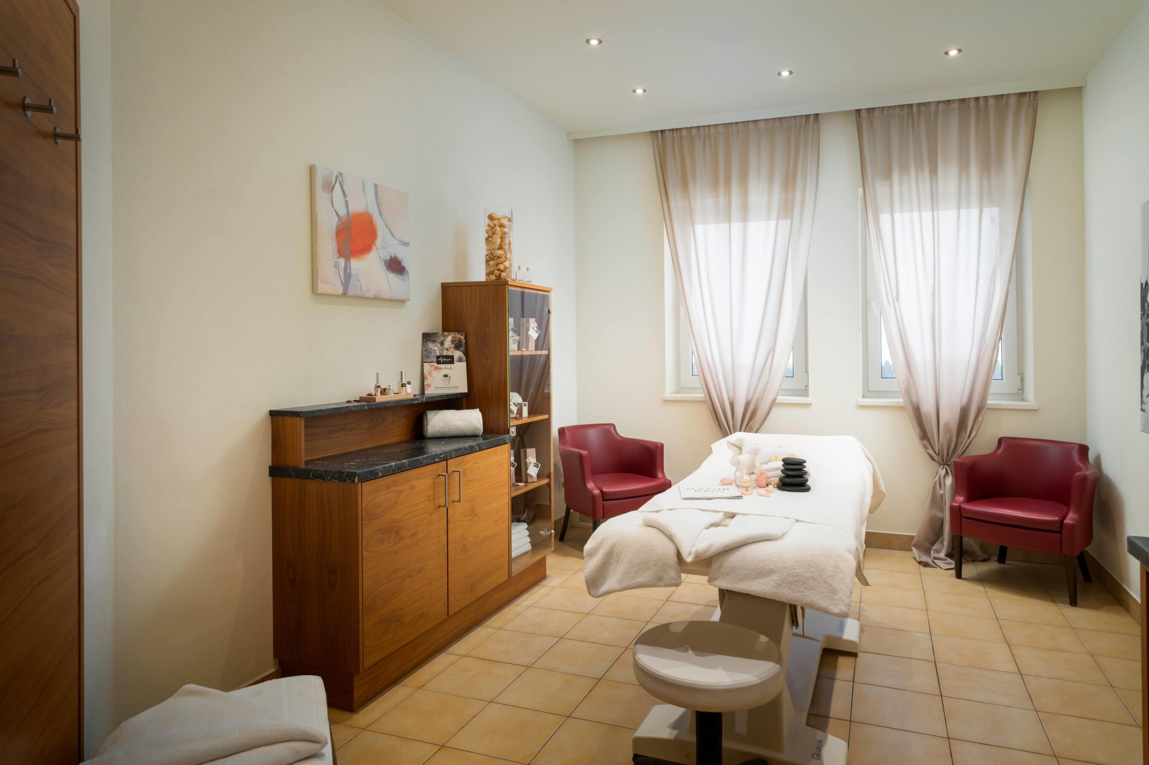 A spa room with a massage table, two red chairs, a wooden cabinet with a glass front, and a large window with curtains.