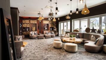 A spacious living area with multiple seating options, including sofas and ottomans, arranged around a central coffee table. The room features a large window providing a view of the outdoors, and the ceiling is adorned with various pendant lights.