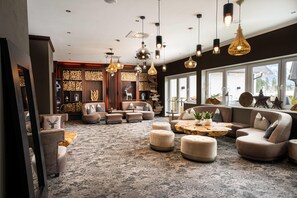 A spacious living area with multiple seating options, including sofas and ottomans, arranged around a central coffee table. The room features a large window providing a view of the outdoors, and the ceiling is adorned with various pendant lights.