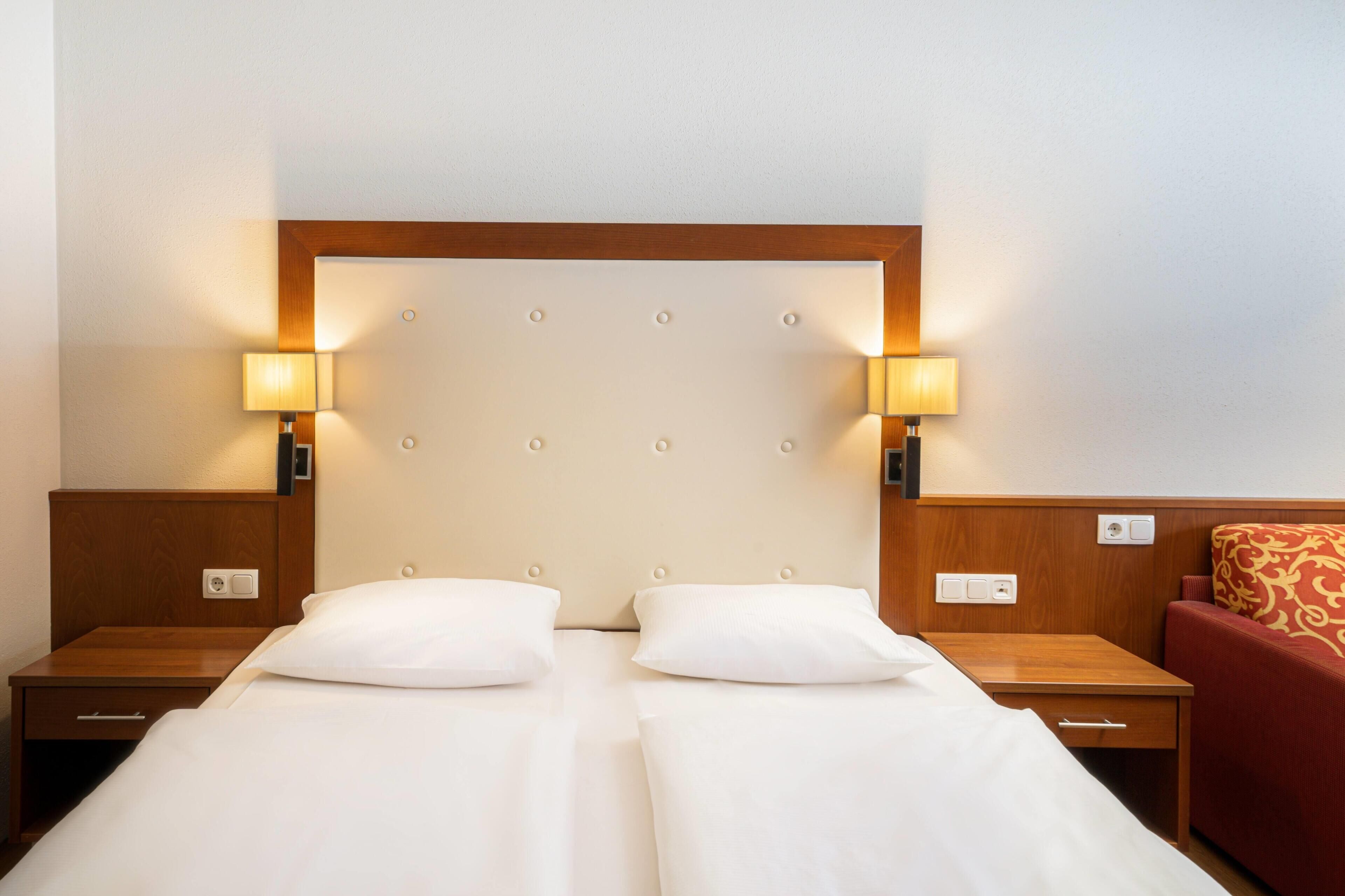 Une chambre d’hôtel avec un lit double, deux tables de chevet, deux lampes fixées au mur et un canapé rouge à motifs.