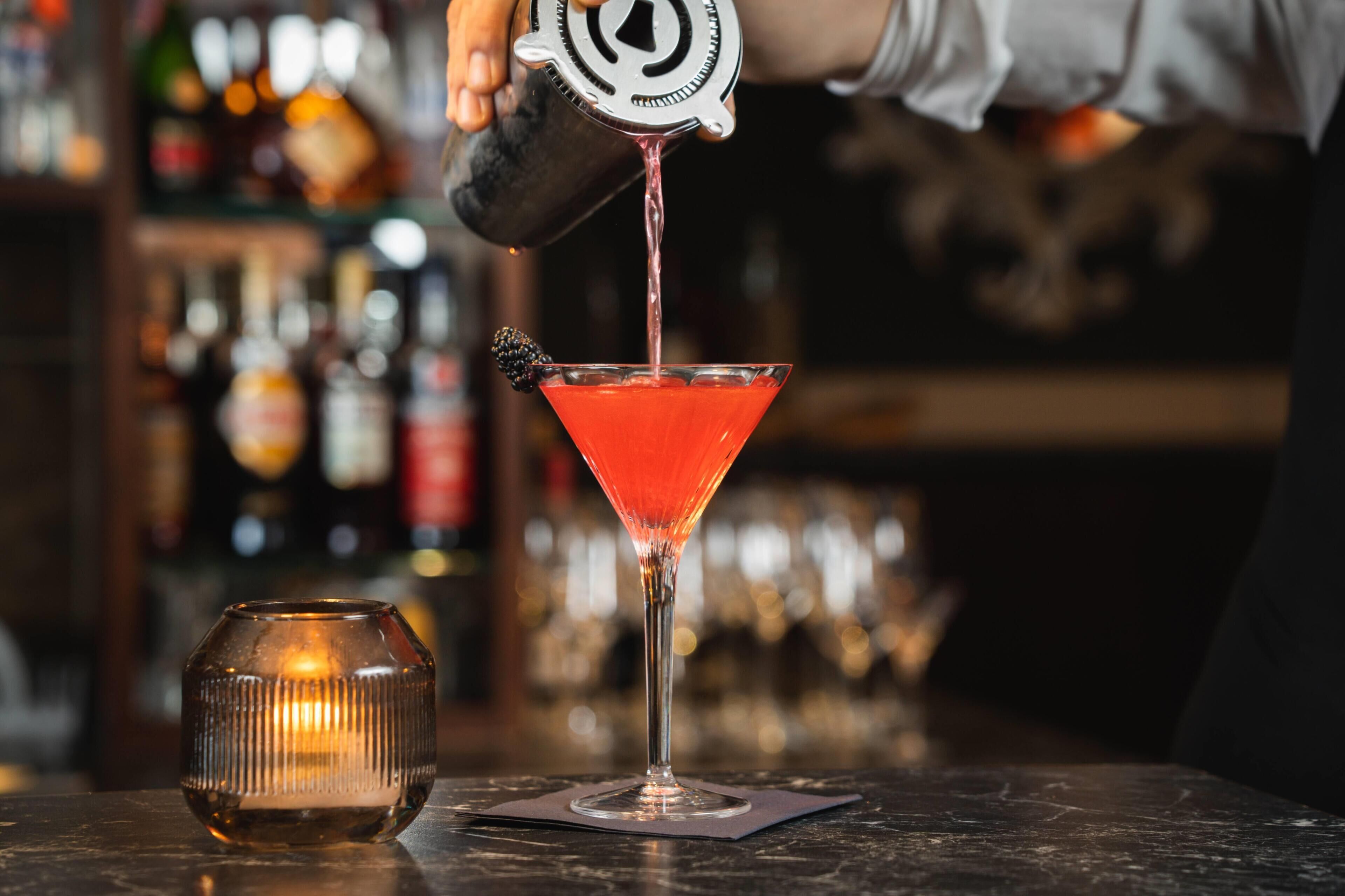 Um bartender servindo um líquido vermelho em uma taça martini, com um enfeite de amora.