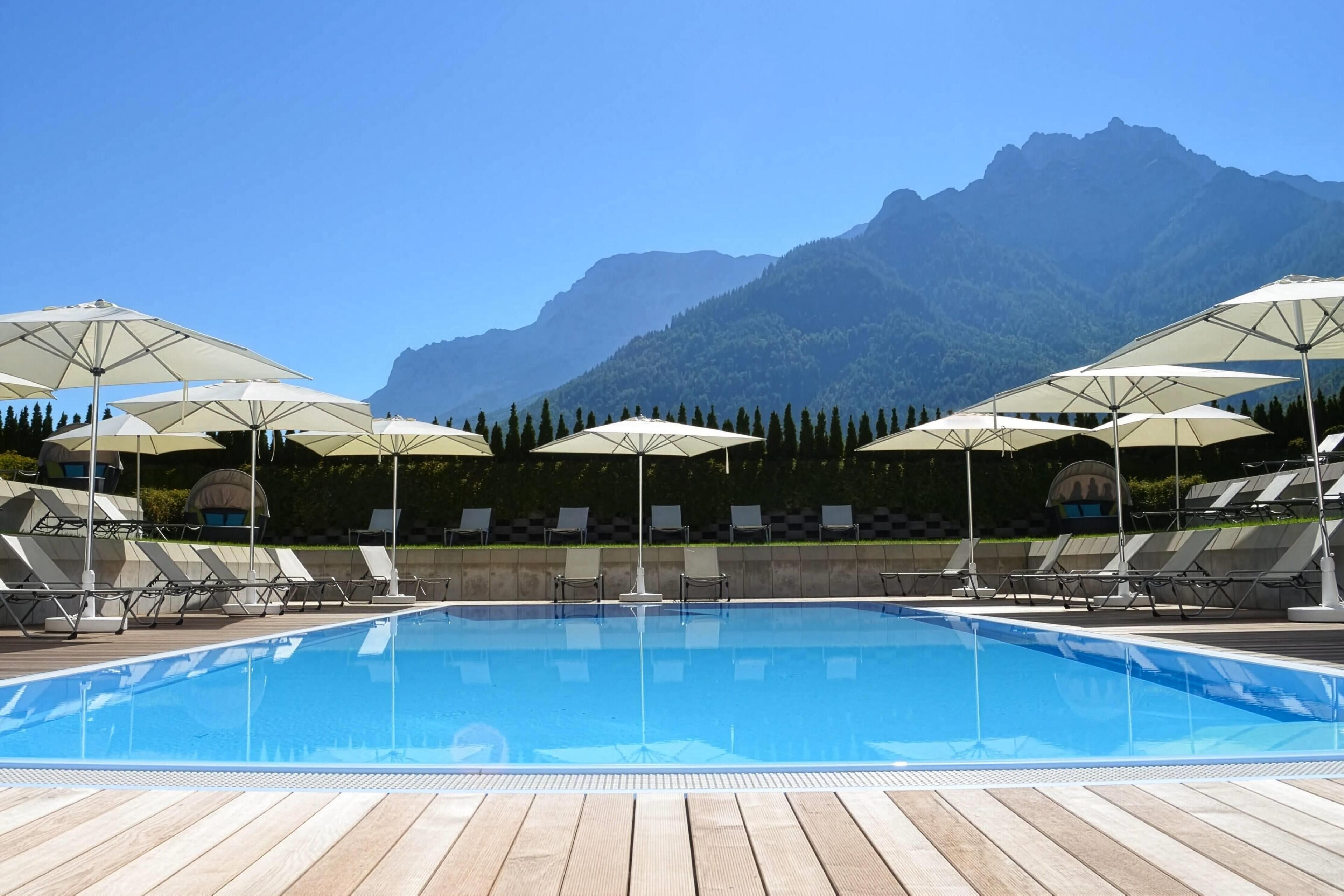 Ein Schwimmbecken mit Liegestühlen und Sonnenschirmen, umgeben von einer Holzterrasse und einem Bergpanorama.