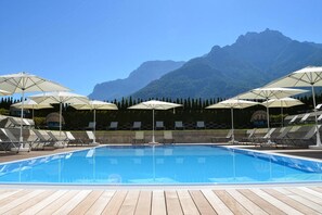 Piscina con sillas de descanso y sombrillas, rodeada por una terraza de madera y un paisaje montañoso.