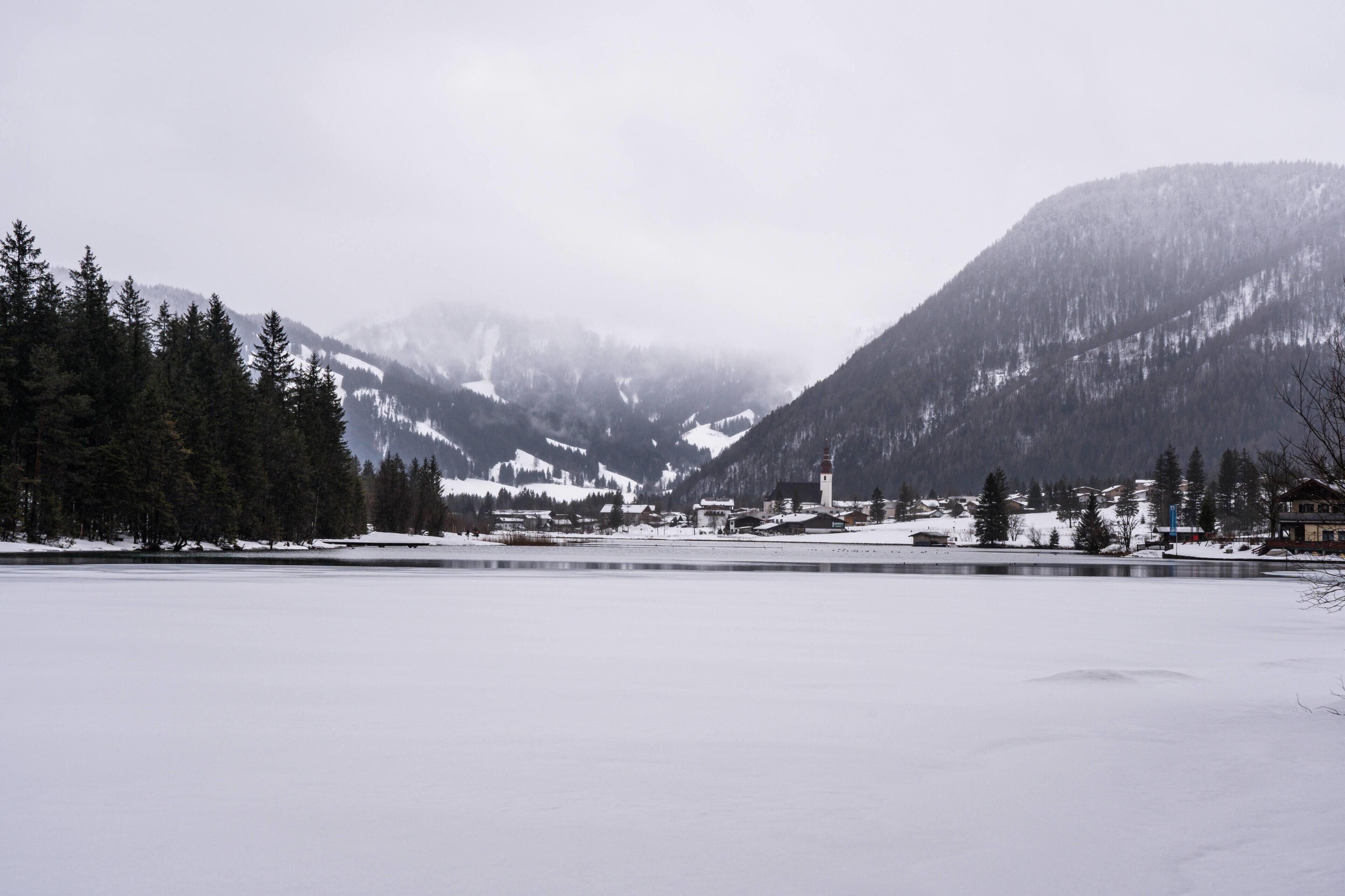 A snow-covered landscape with a lake, mountains, and a small settlement with buildings.