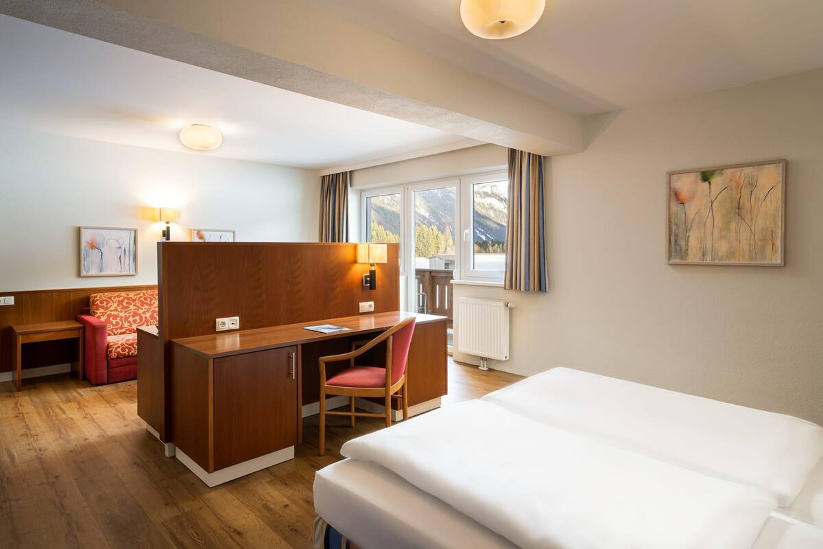 Une chambre d’hôtel avec un bureau en bois, une chaise rouge, un lit avec du linge de maison blanc, une fenêtre donnant sur les montagnes et un tableau accroché au mur.