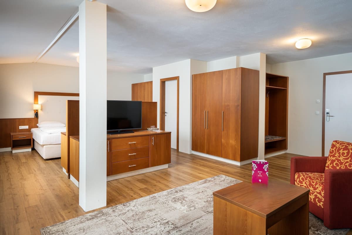 Une chambre d’hôtel moderne avec un plancher en bois, une grande armoire, une télévision, un lit, un canapé rouge à motifs et une petite table sur laquelle se trouve un sac rose.