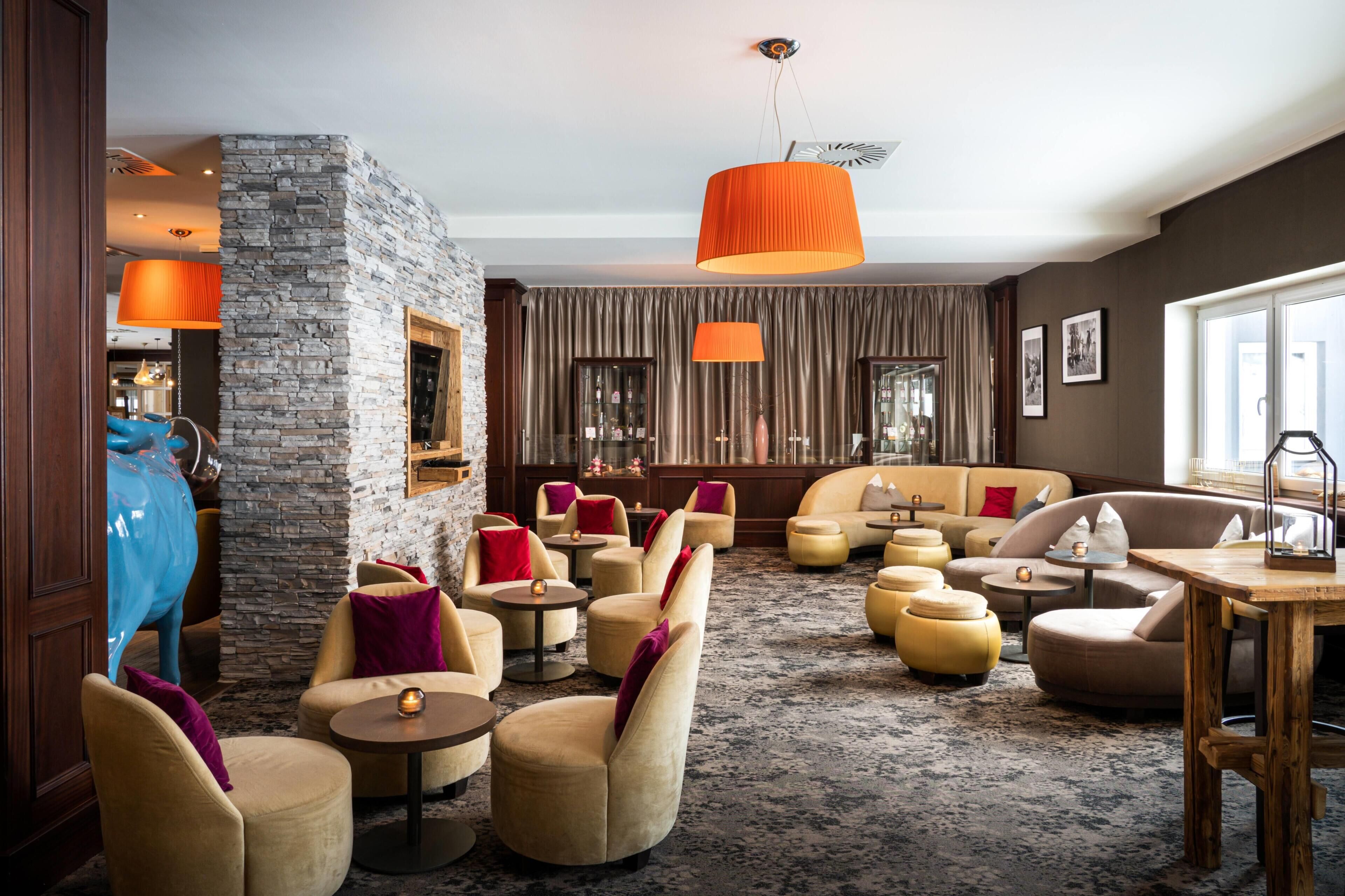 Un espacio lounge moderno con sofás, sillones y mesas redondas, destacando una pared de piedra y una lámpara colgante naranja de gran tamaño.