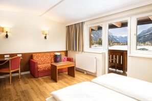 Une chambre d’hôtel avec un plancher en bois, un canapé rouge, une petite table, un lit blanc et une fenêtre donnant sur la montagne.