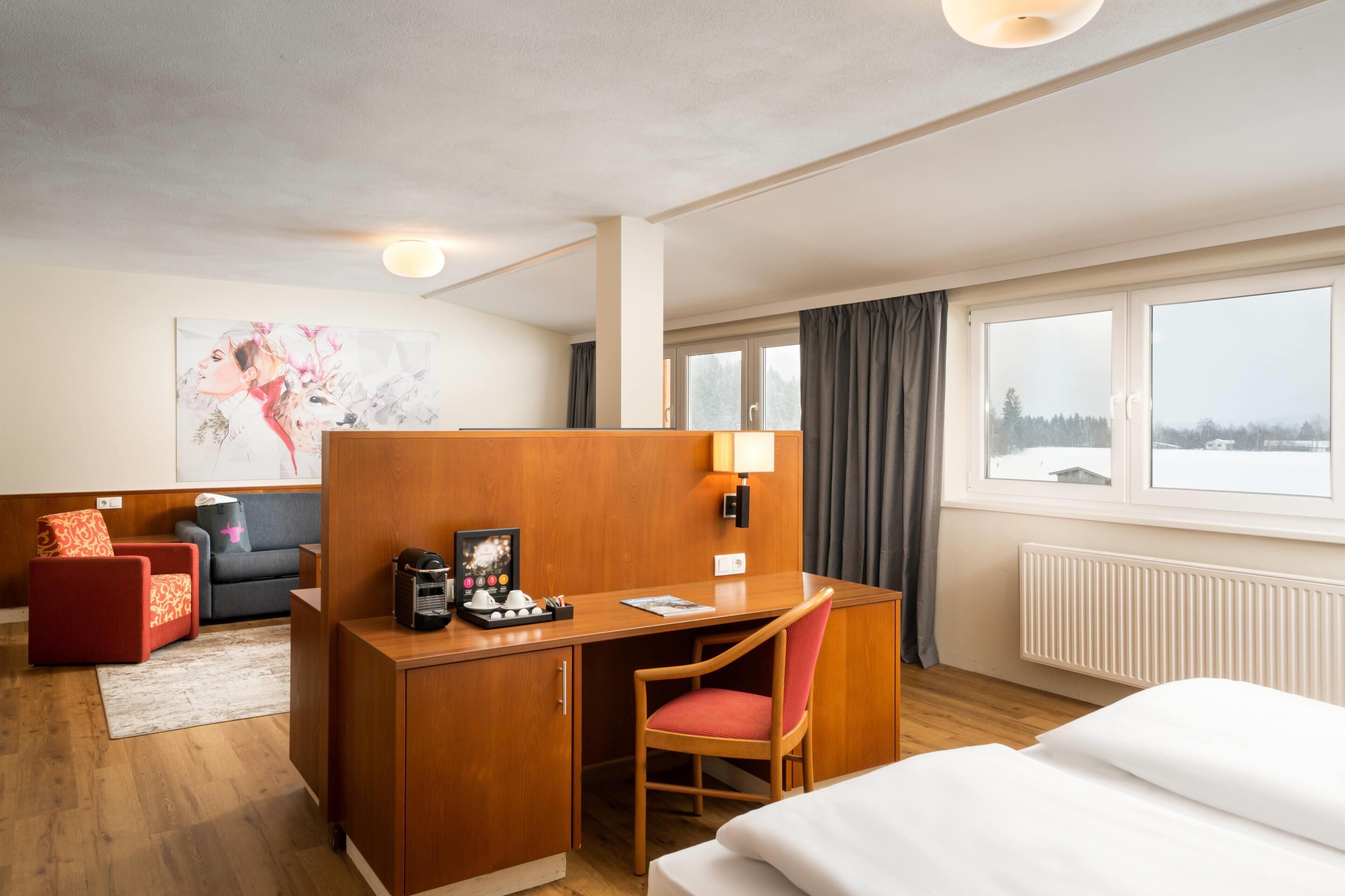A hotel room with a wooden desk, a red chair, a bed, a sofa, and a large window with a view.