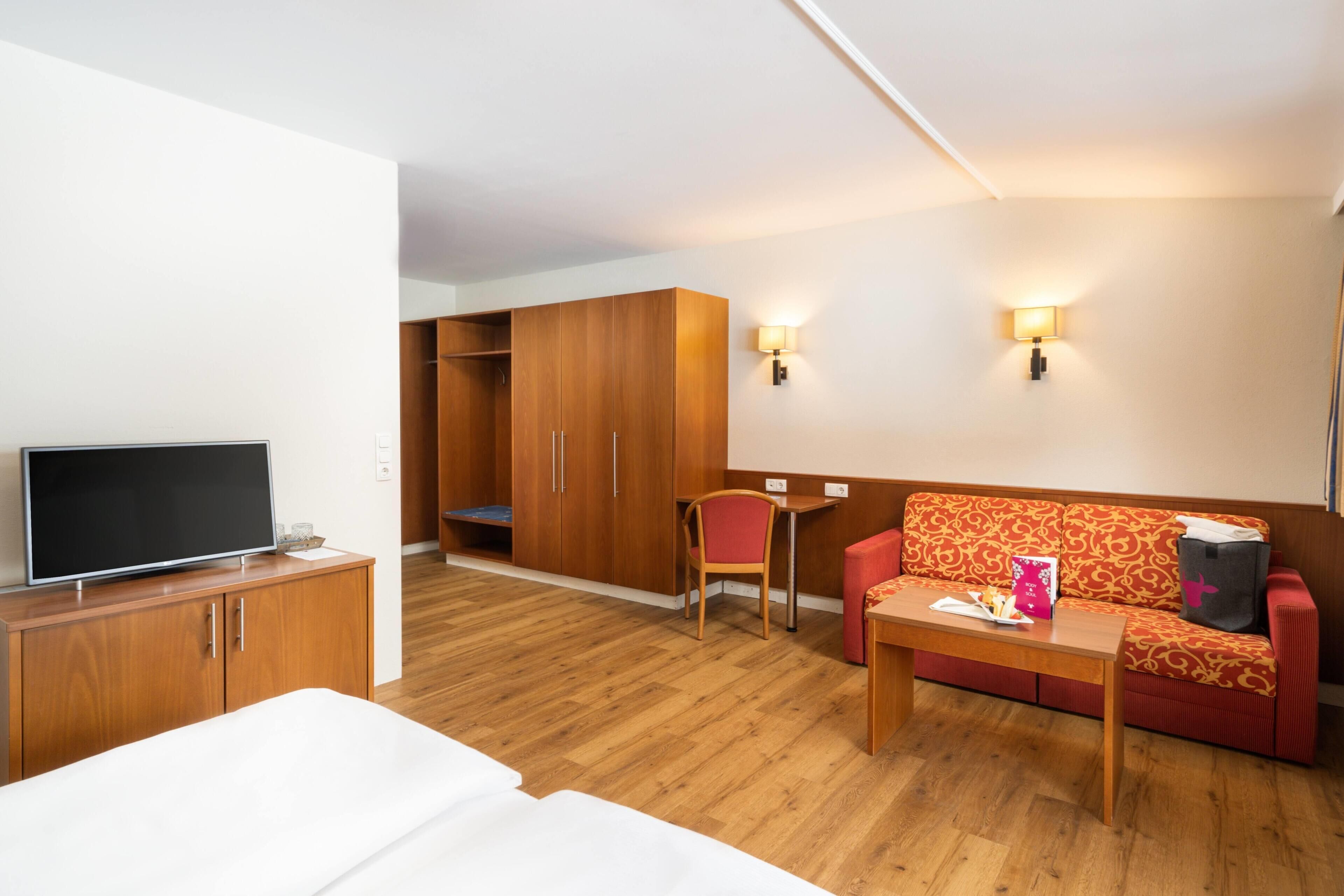 Une chambre d’hôtel avec un lit, une télévision, une armoire en bois, un canapé rouge, une petite table et deux lampes fixées au mur.