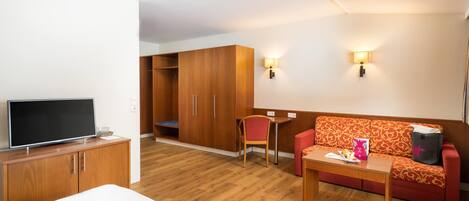 A hotel room with a bed, a TV, a wooden wardrobe, a red sofa, a small table, and two wall-mounted lamps.