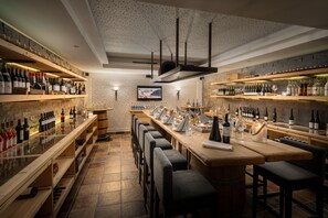 A bar area with multiple shelves of bottles, a long counter with bar stools, and a television screen on the wall.
