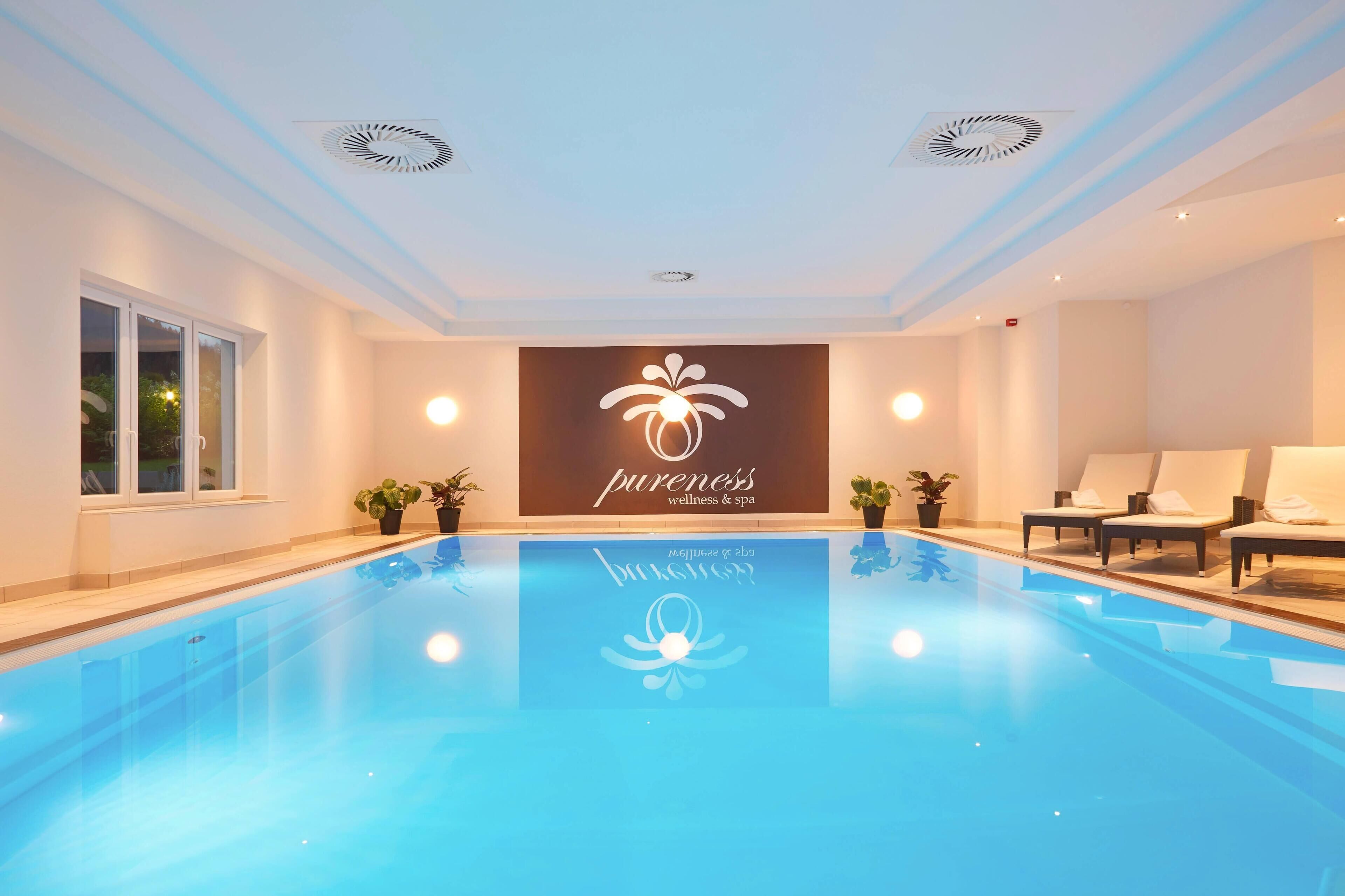 A modern indoor pool area with a clear blue pool, lounge chairs, and a wall with a wellness spa logo.