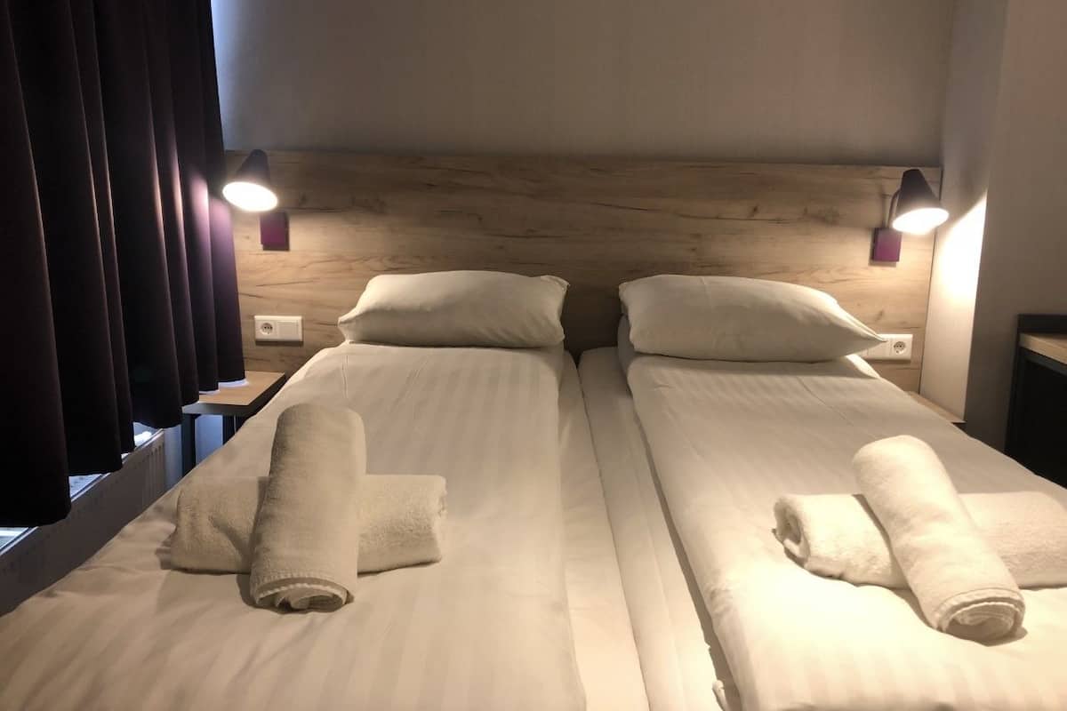 A double bed with two pillows and two rolled towels, a wooden headboard, and two bedside lamps.